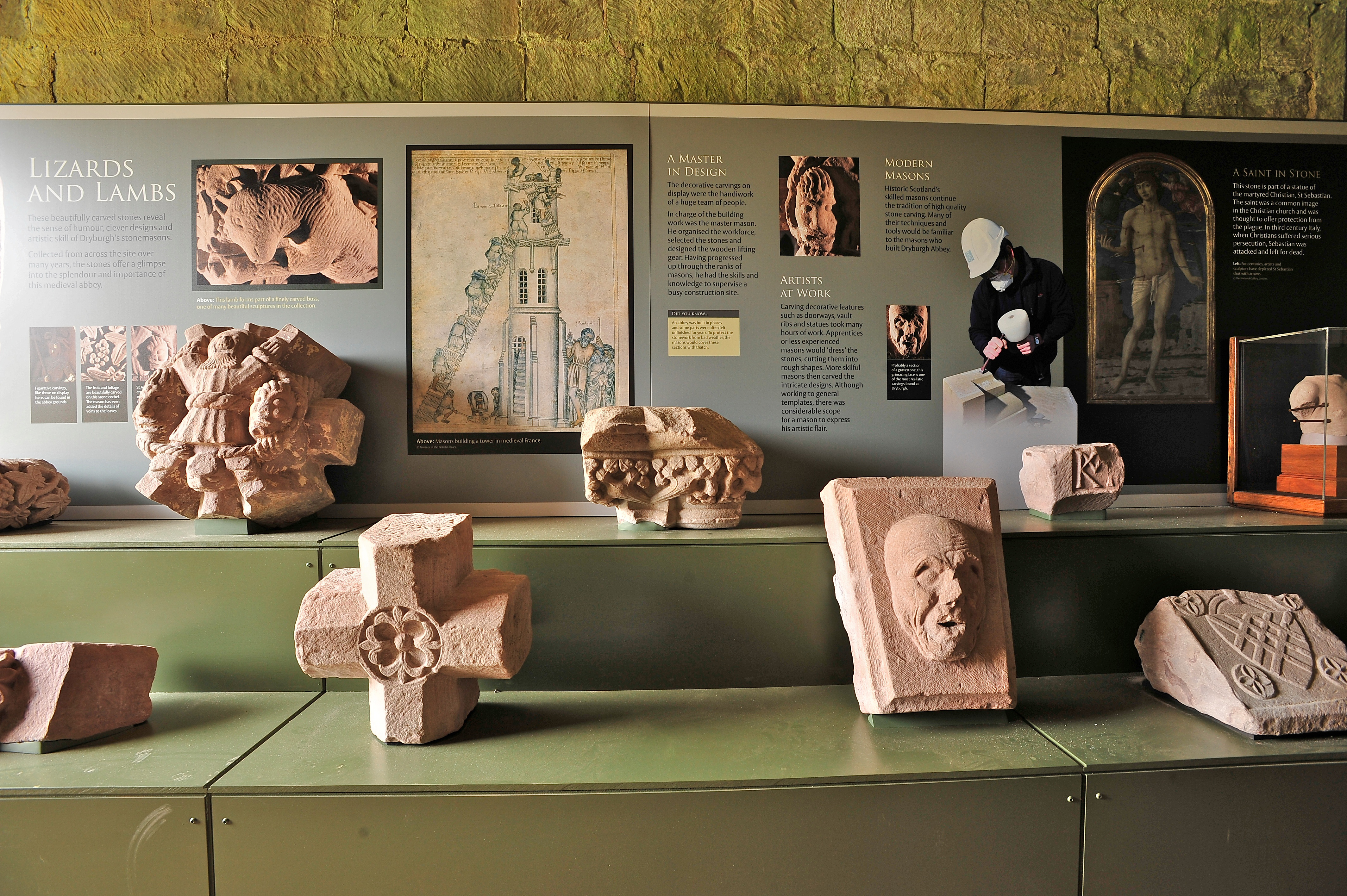 An exhibit in a museum. Stone carvings sit on two platforms at different heights. Carvings include a face carving and a cross. There is information on a panel behind them, with the title 'LIZARD AND LAMBS' visible. 
