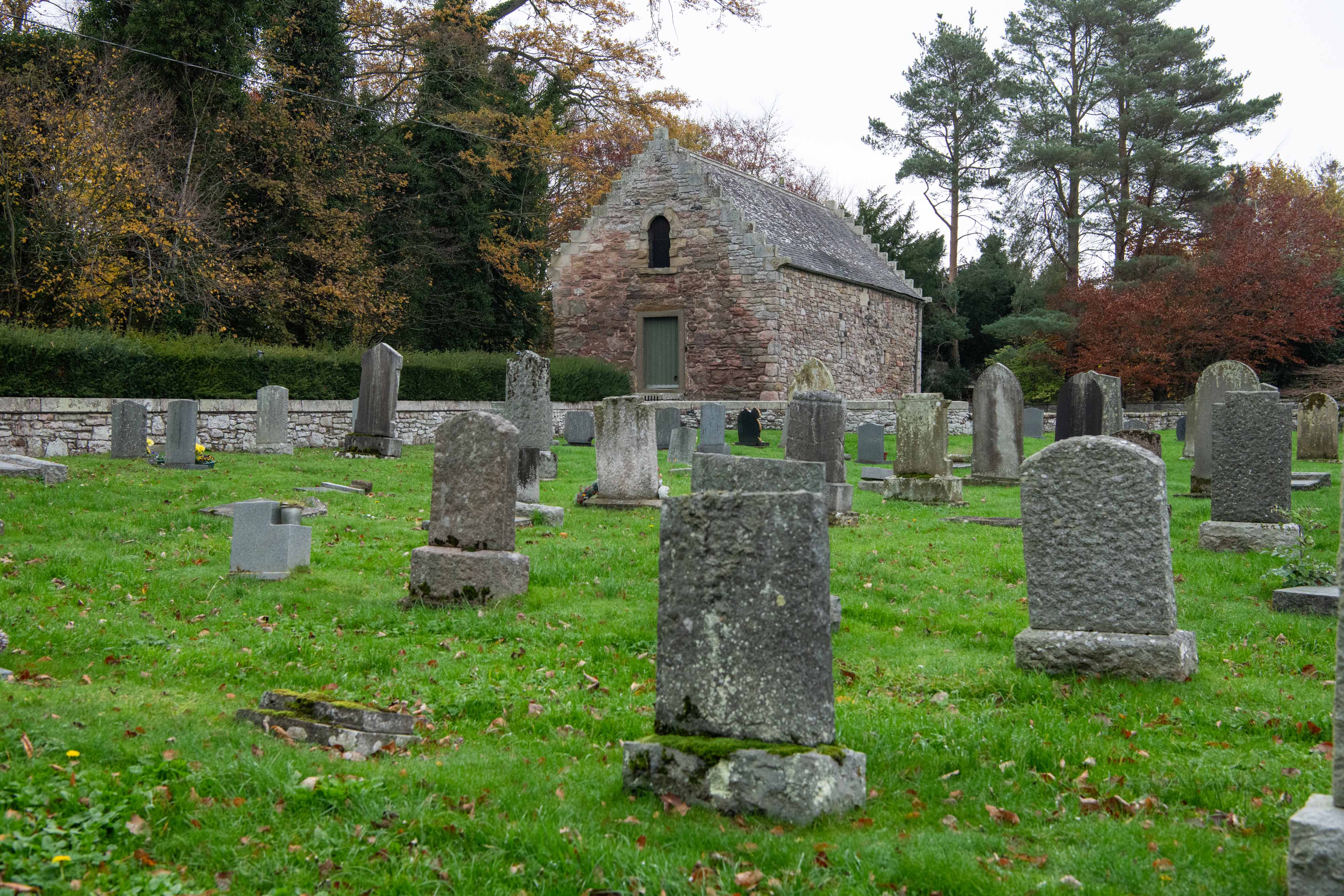 Many historic gravestones in front of a sandstone barn