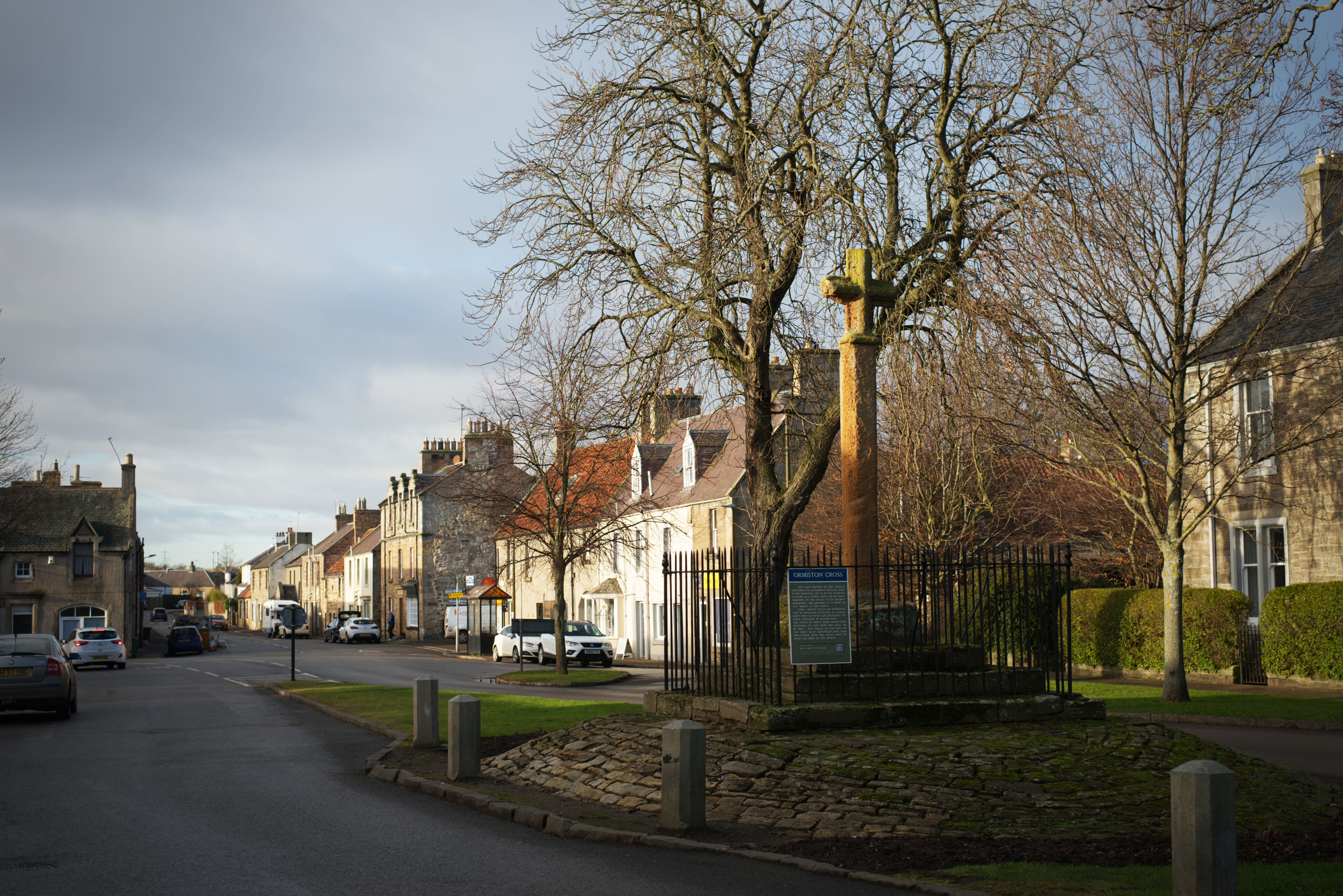 View of Ormiston Market Cross standing on a raised circular stone platform and enclosed by a black iron fence. The tall, weathered stone shaft with a small carved cross at the top is set beside leafless trees on a village street. Rows of stone houses line the road to the left, with parked cars and soft afternoon light illuminating the scene.