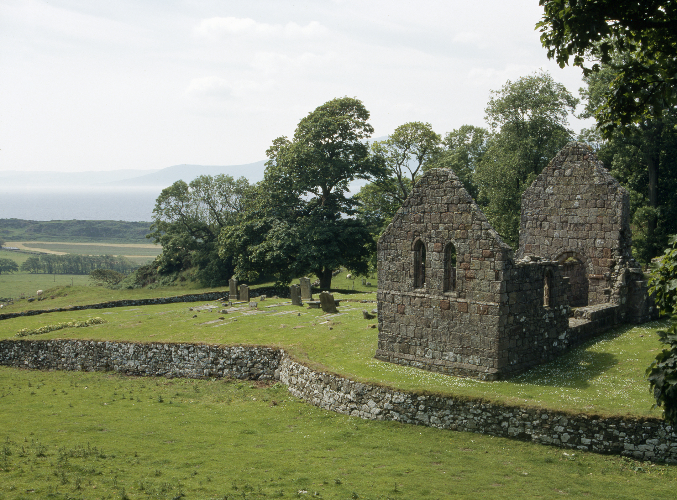 Ruins of St Blane’s Church on a grassy hillside, featuring two tall gable ends with narrow arched windows and partially collapsed stone walls. A low stone boundary wall encloses the site, with several upright gravestones scattered nearby. Behind the church, dense green trees frame the view, and in the distance, the sea and faint outlines of hills are visible under a pale sky.