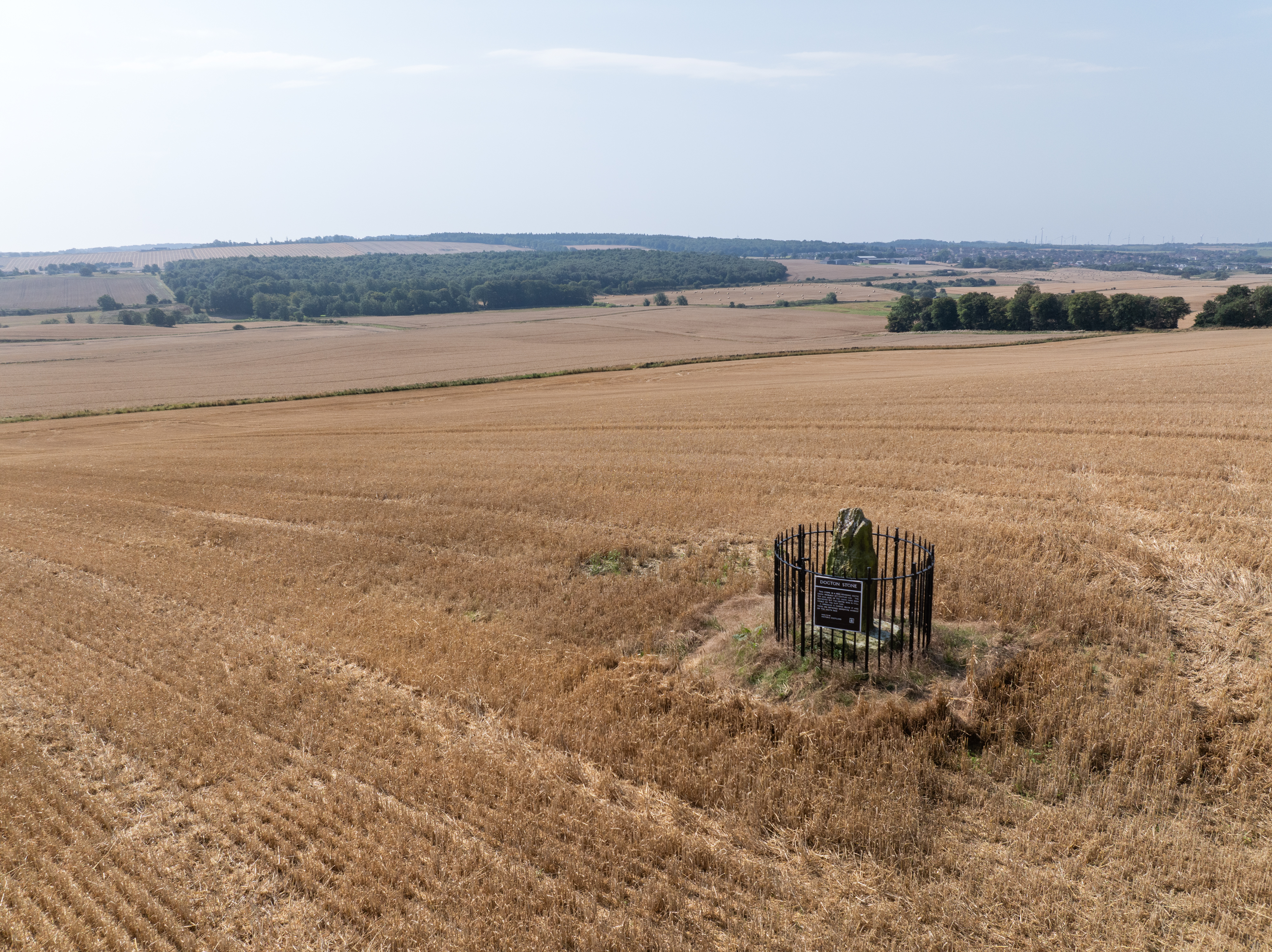 An aerial view of the Dogton Stone enclosure standing alone in the centre of a large, recently harvested field. The landscape extends to distant woodland and farmland.