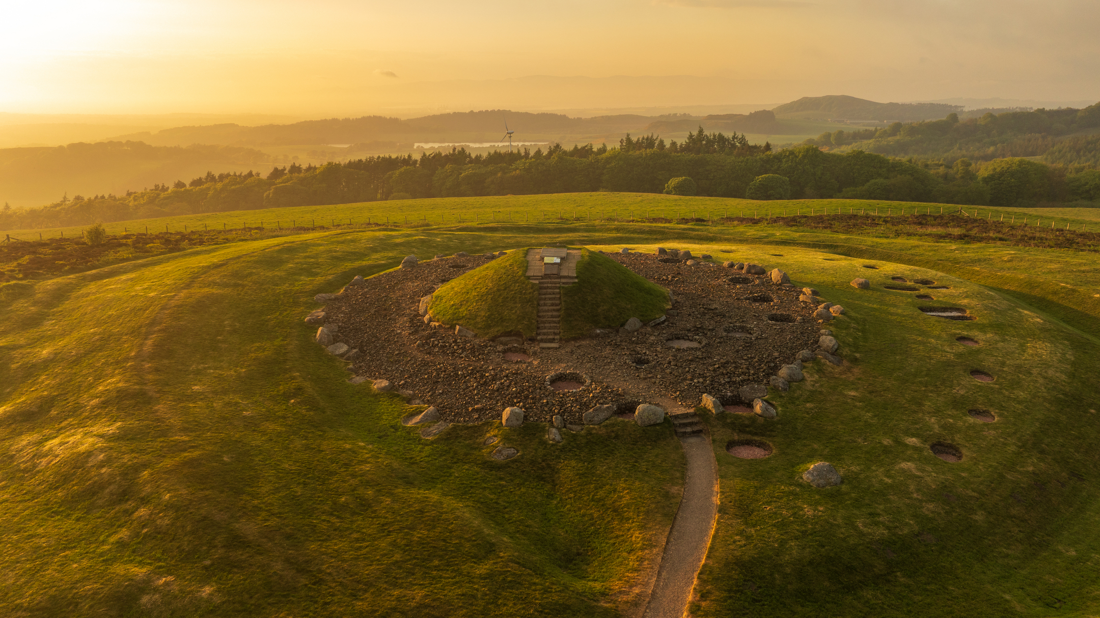 Golden sunlight across a luscious green landscape with a circular area with stones, holes that have been dug in circular shapes and a mound with a staircase on it leading to an entrance