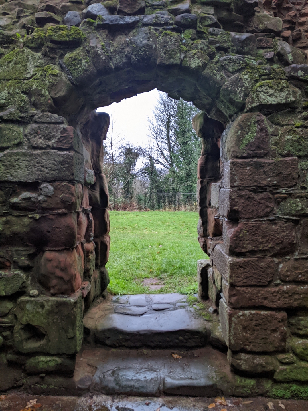 A stone doorway, which shows grass and trees on the other side. There is a small step, the stones have a dusting of moss on them.