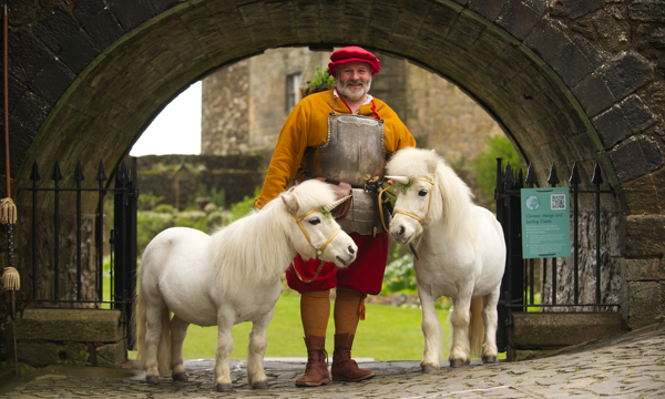 Man in medieval dress stands in a stone archway. Two small white ponies with unicorn horns stand on either side of him.