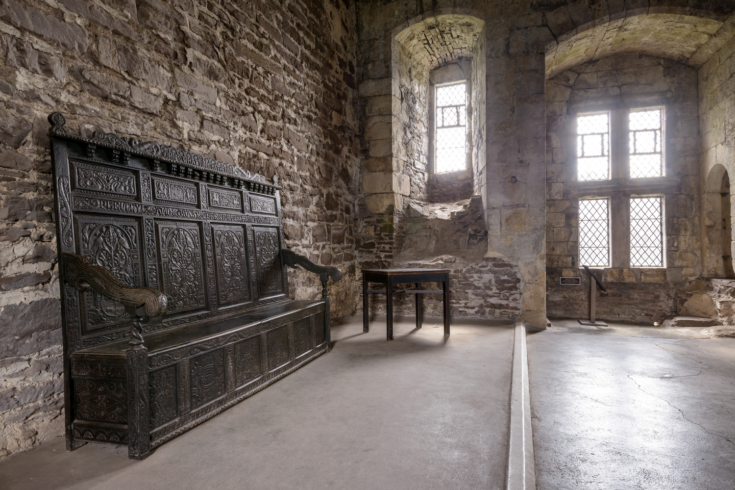 A historical stone building, with two large windows with arches above them. There is a long wood seat with detailed engravings leaning against the wall. There is a table nearby.