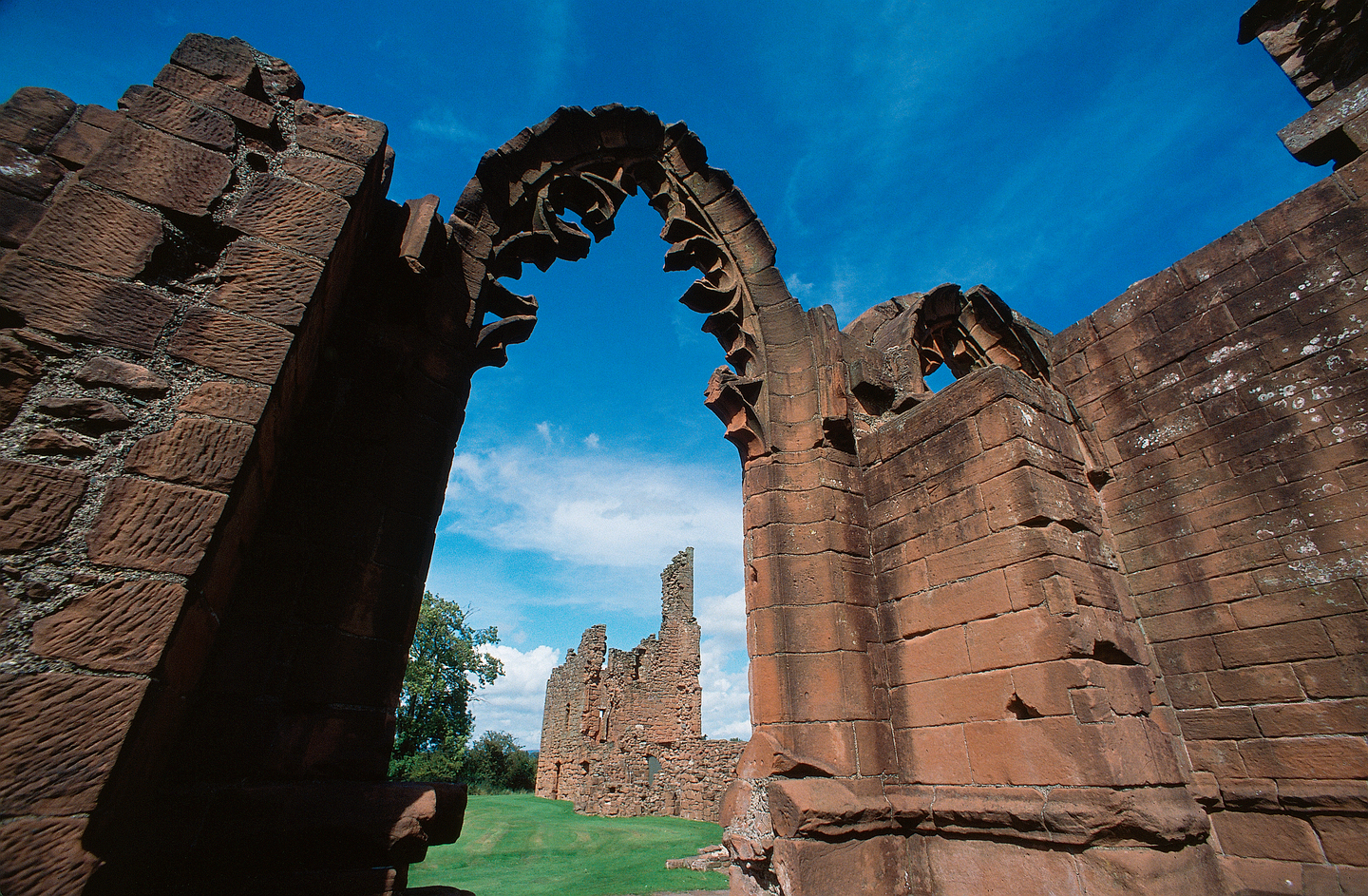 A large stone arch, attached to a wall either side. There is detailing around the inside of the arch, and you can see wall remains in the background through the arch. It is a sunny day.