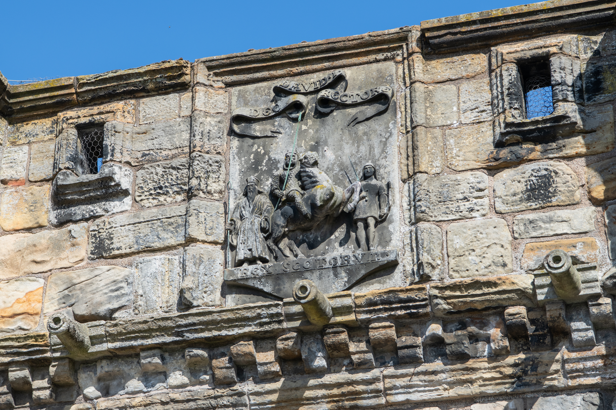 Detailed close-up of the carved stone panel above the main arch of the West Port. The relief depicts several figures surrounding a central mounted rider, with decorative banners and inscriptions above and below. The stone surface shows signs of age and weathering, set within the surrounding ashlar masonry.