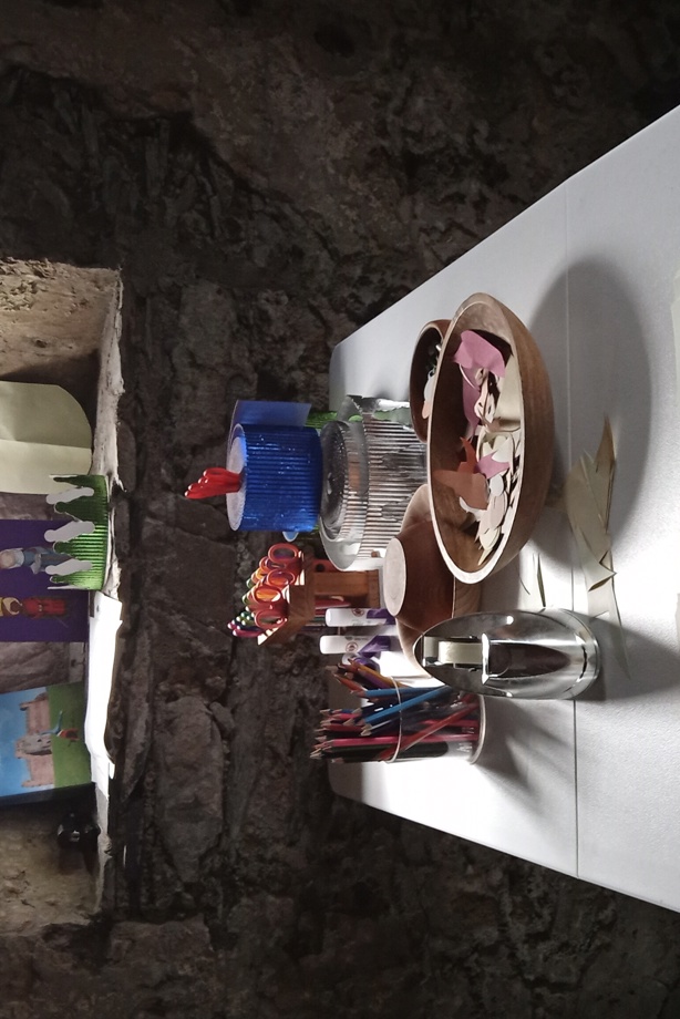 Various arts and crafts materials (coloured pencils, scissors, sellotape, paper, craft instructions and example crafted crown and shield) laid out on some long tables in a castle room.