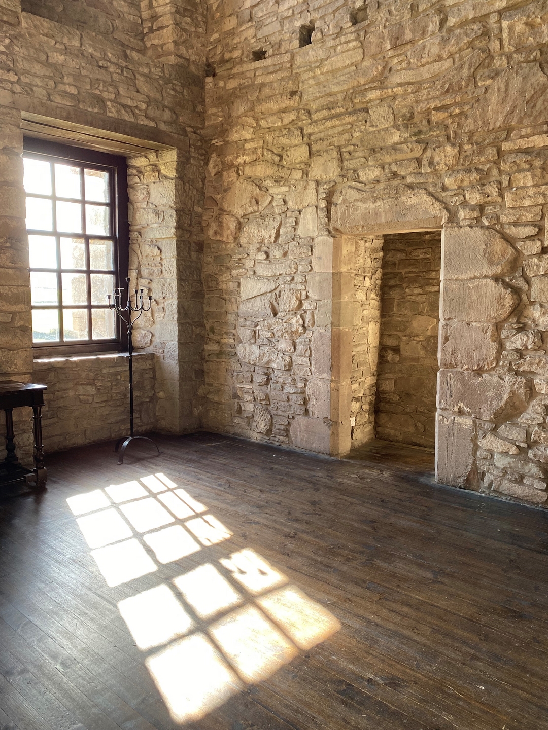 Interior of a stone building, with a large window with light coming though, reflecting onto the floor. There is an exit in the wall, due to there being a corner behind it, you can only see stone. 