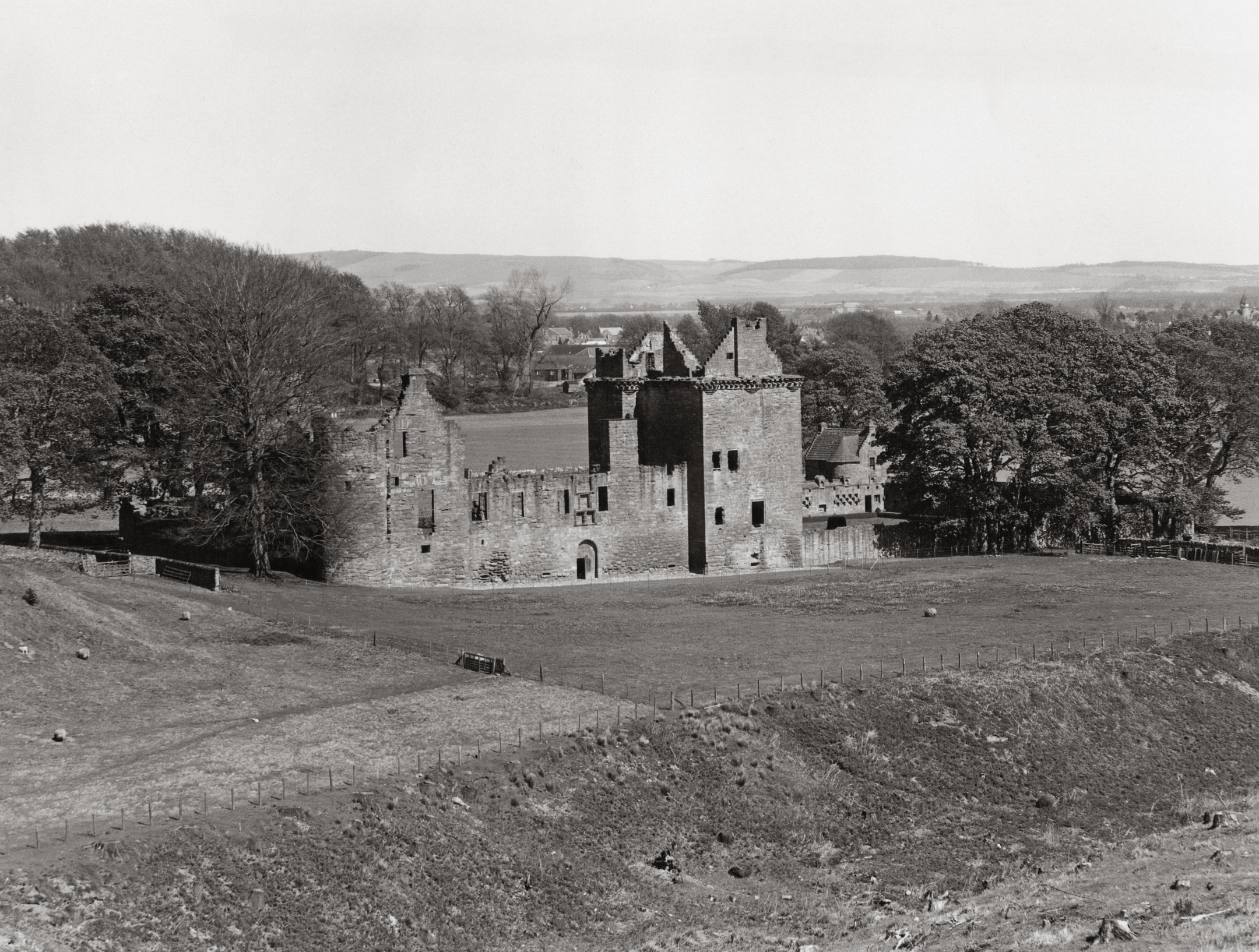 A black and white archive image showing a ruined castle surrounded by trees and farmland. 