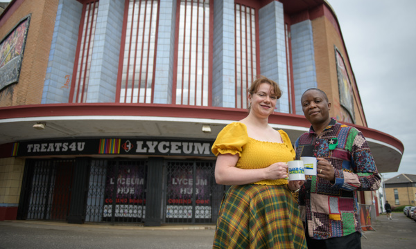 Two people stand in front of the entrance to the Lyceum Theatre, they are facing the camera posing for a photo while holding white mugs with Talking about Heritage written on them in logo format.