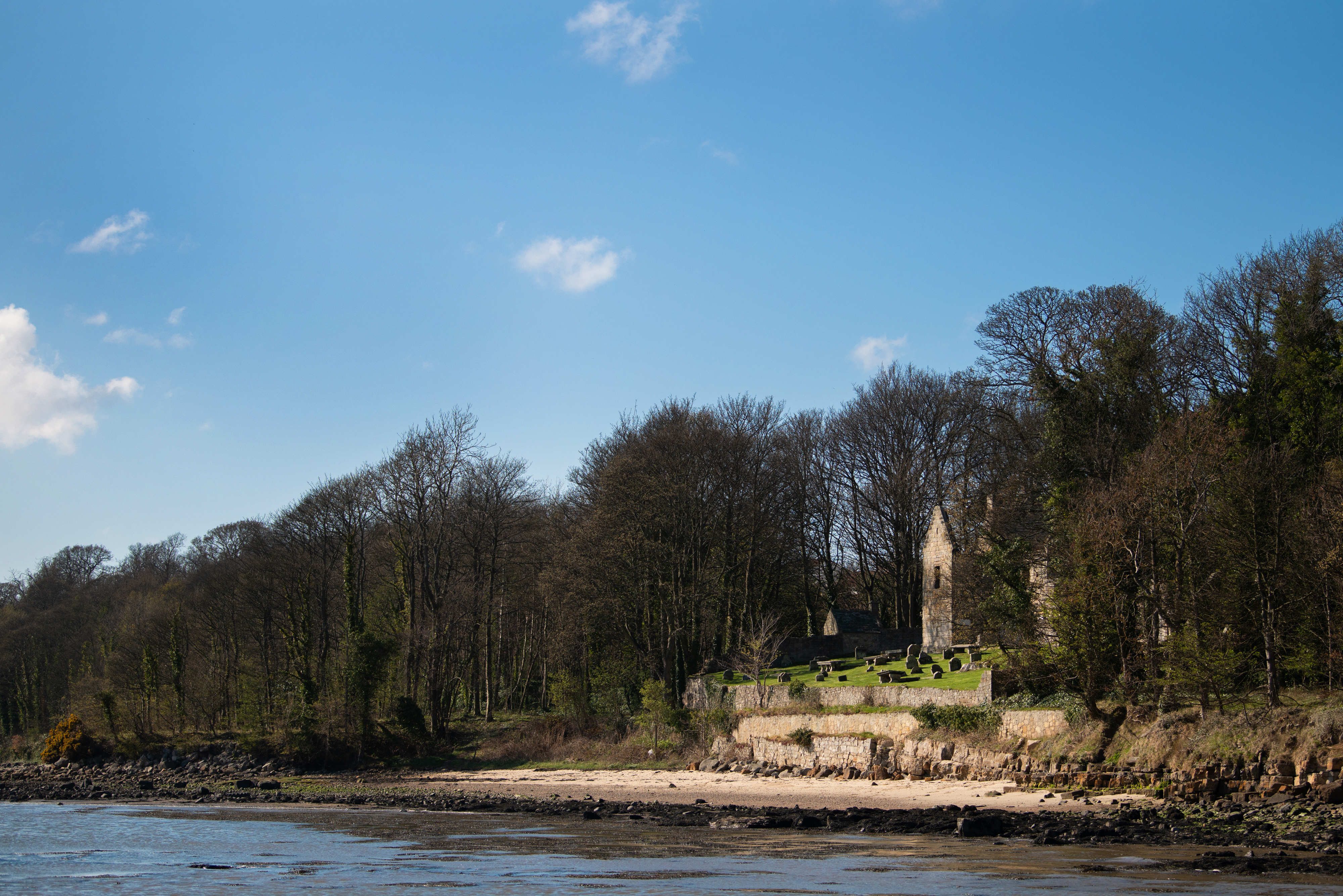 View from the shoreline looking toward St Bridget’s Kirk, with the ruins and graveyard partly hidden by trees above the beach.