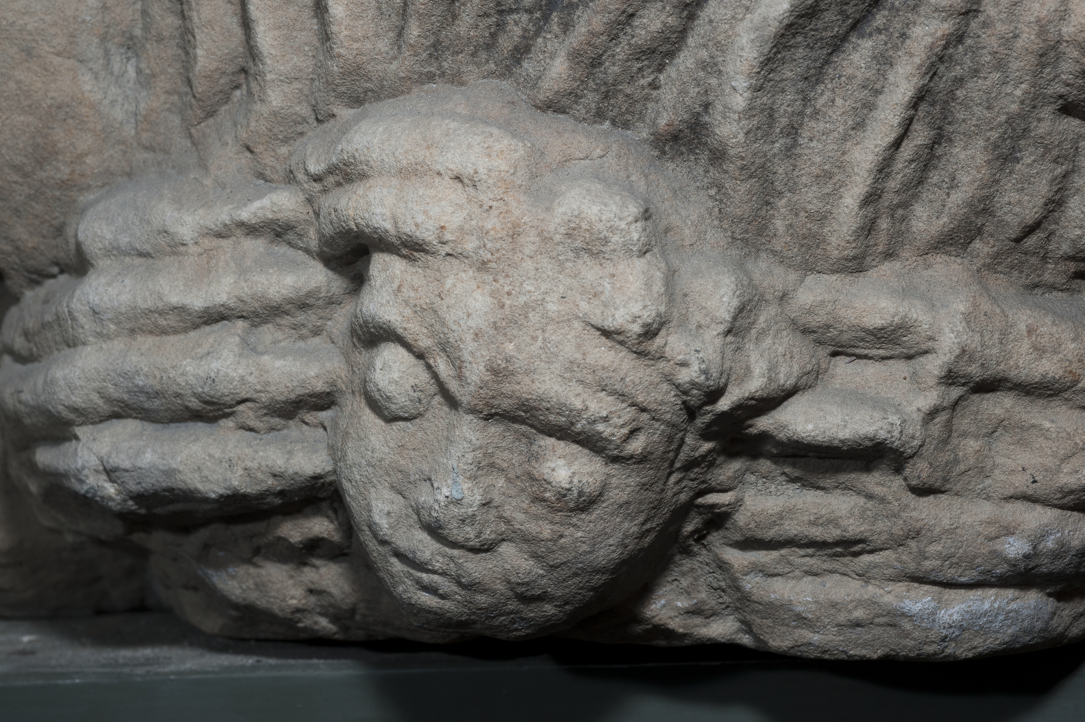 A close-up of a carved stone fragment showing a small, rounded human-like face emerging from sculpted folds of stone. The surface is worn and textured, highlighting the age of the medieval carving.