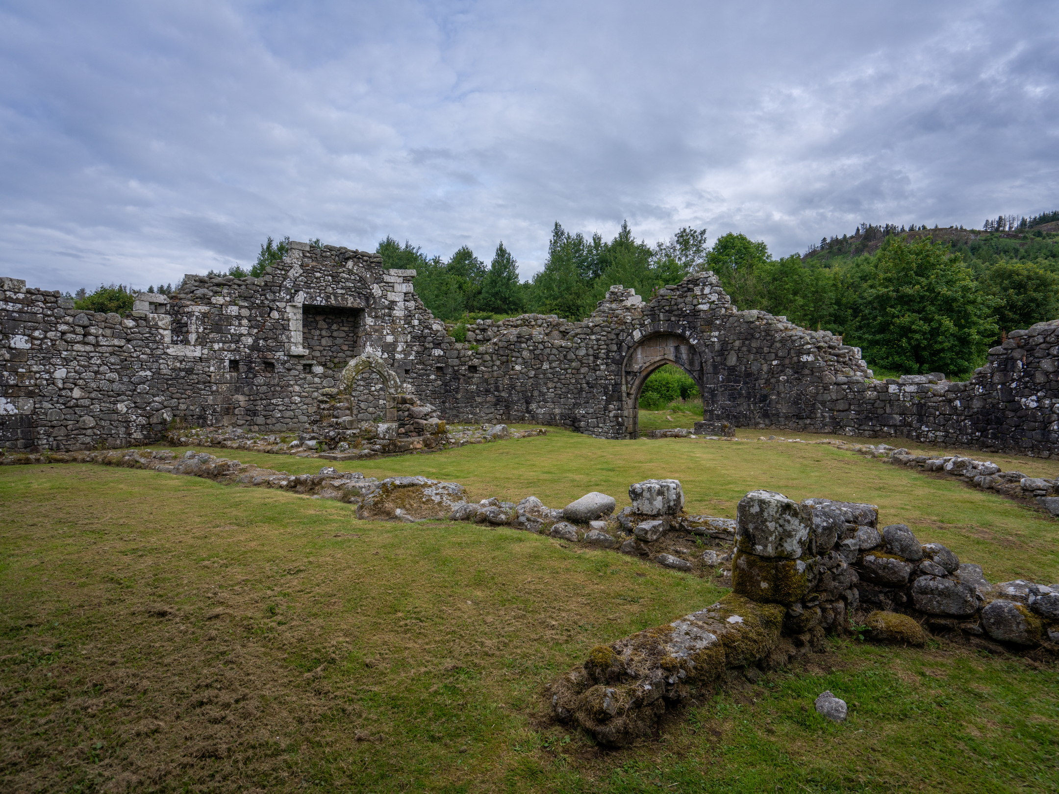 A wide interior view of Loch Doon Castle’s stone foundations and surviving walls, featuring an arched doorway set against surrounding greenery and an overcast sky.