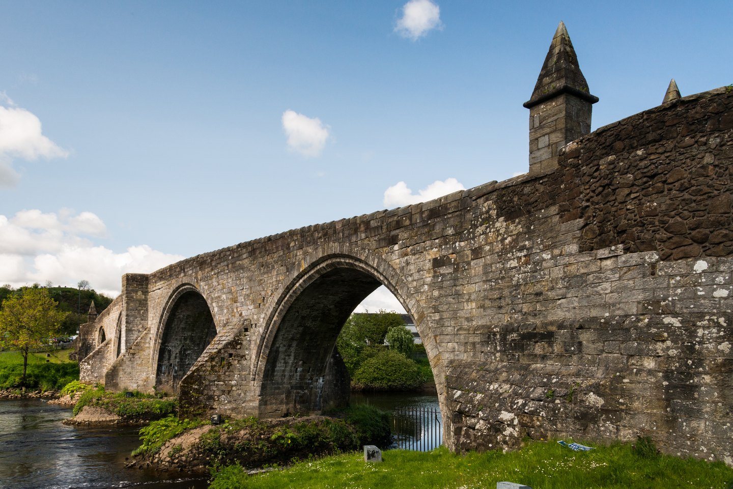 Side view of Stirling Old Bridge showing its stone arches spanning the River Forth under a blue sky.