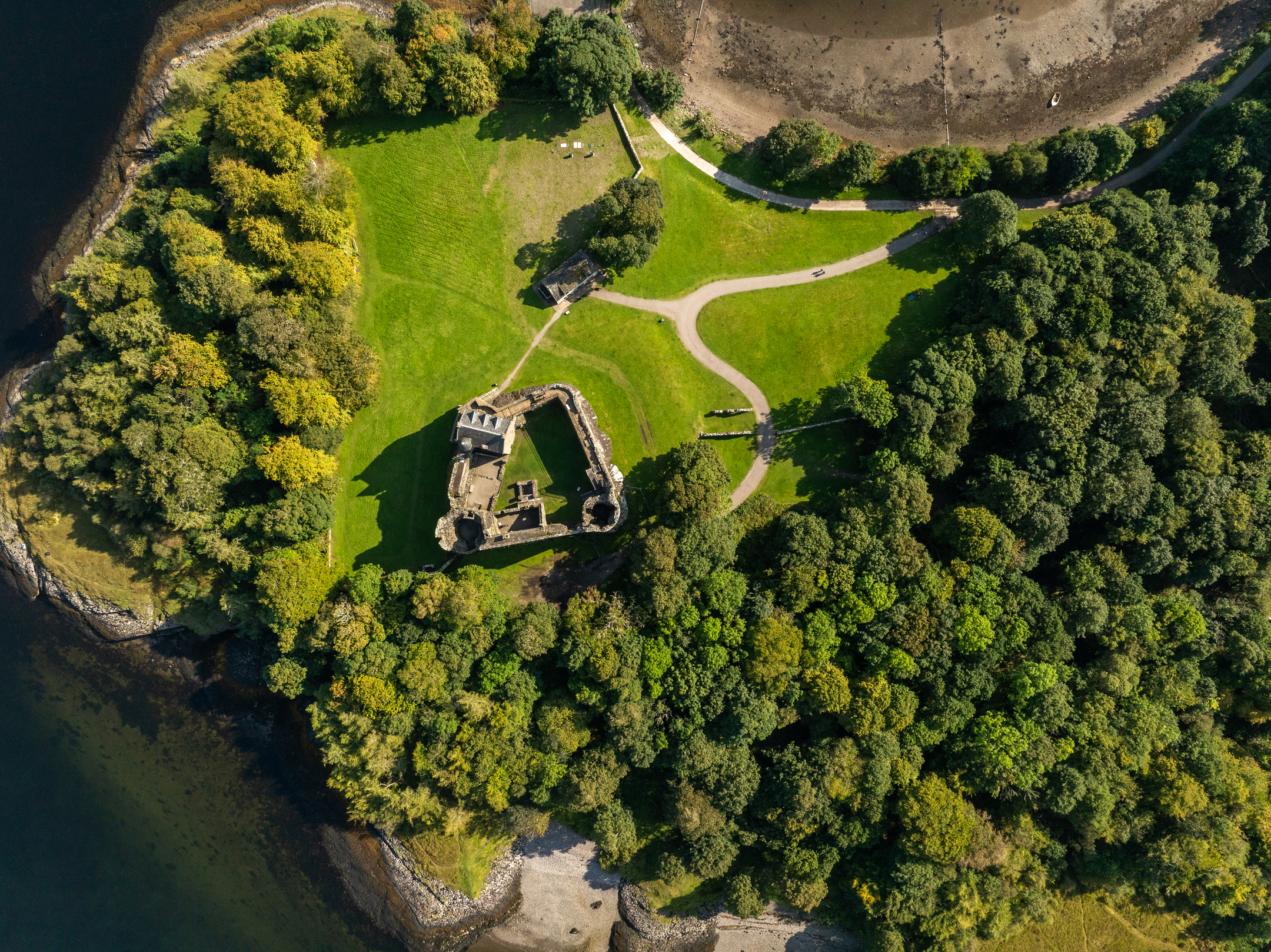 Aerial view of a peninsula with a castle ruin at its centre, surrounded by manicured lawns and dense woodland.