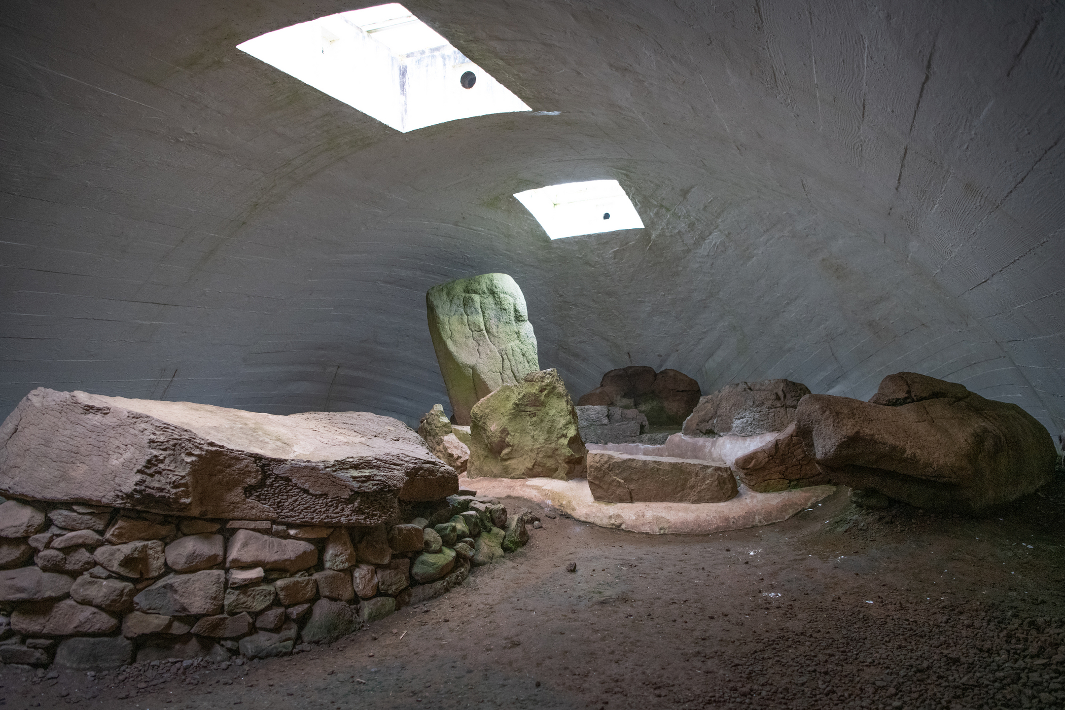 Large stones, and a large stone on top of smaller stones, inside some sort of bunker with two windows or entrances at the top with light shining in