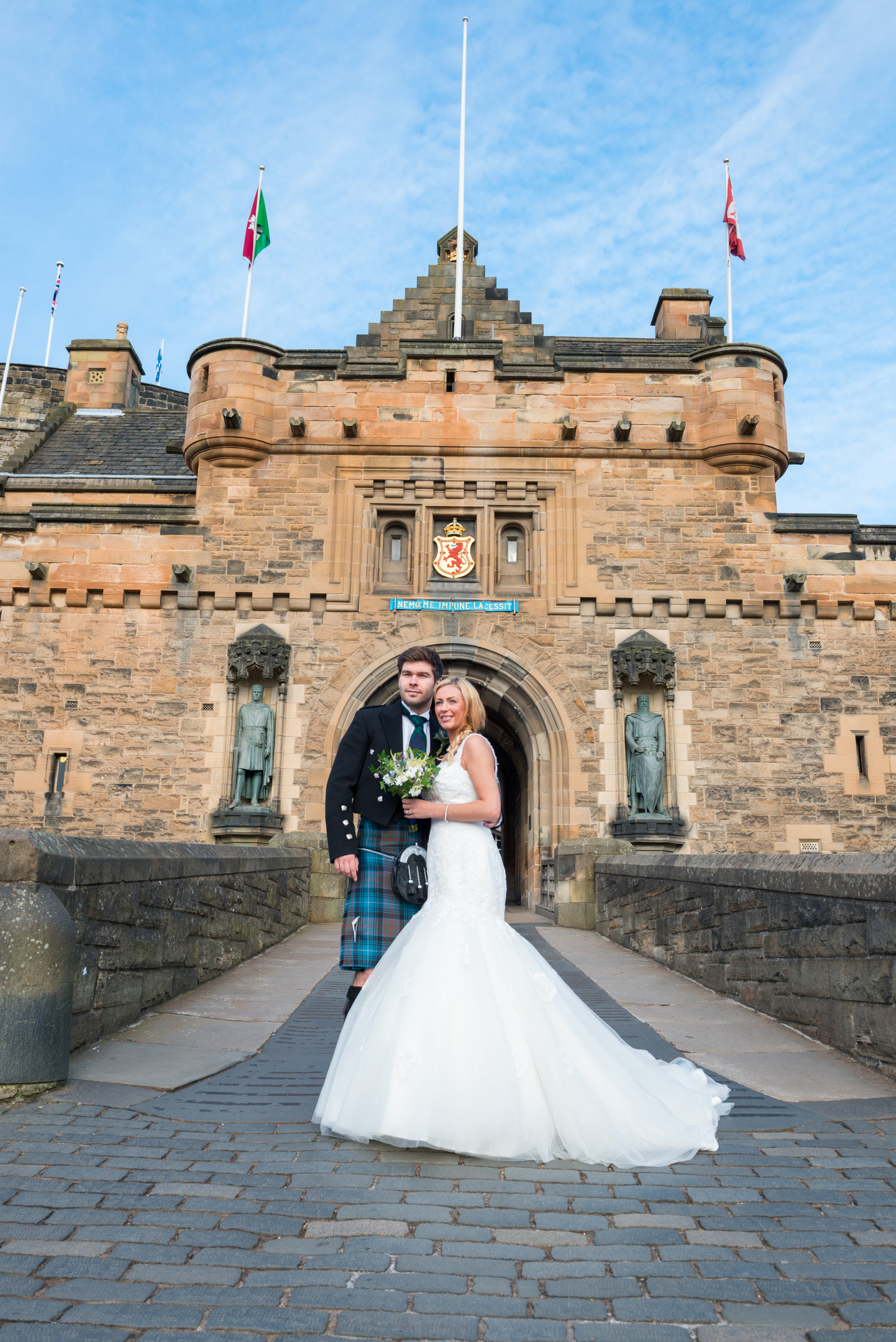 Two adults in formal wedding attire standing on a stone bridge in front of a historic castle entrance.