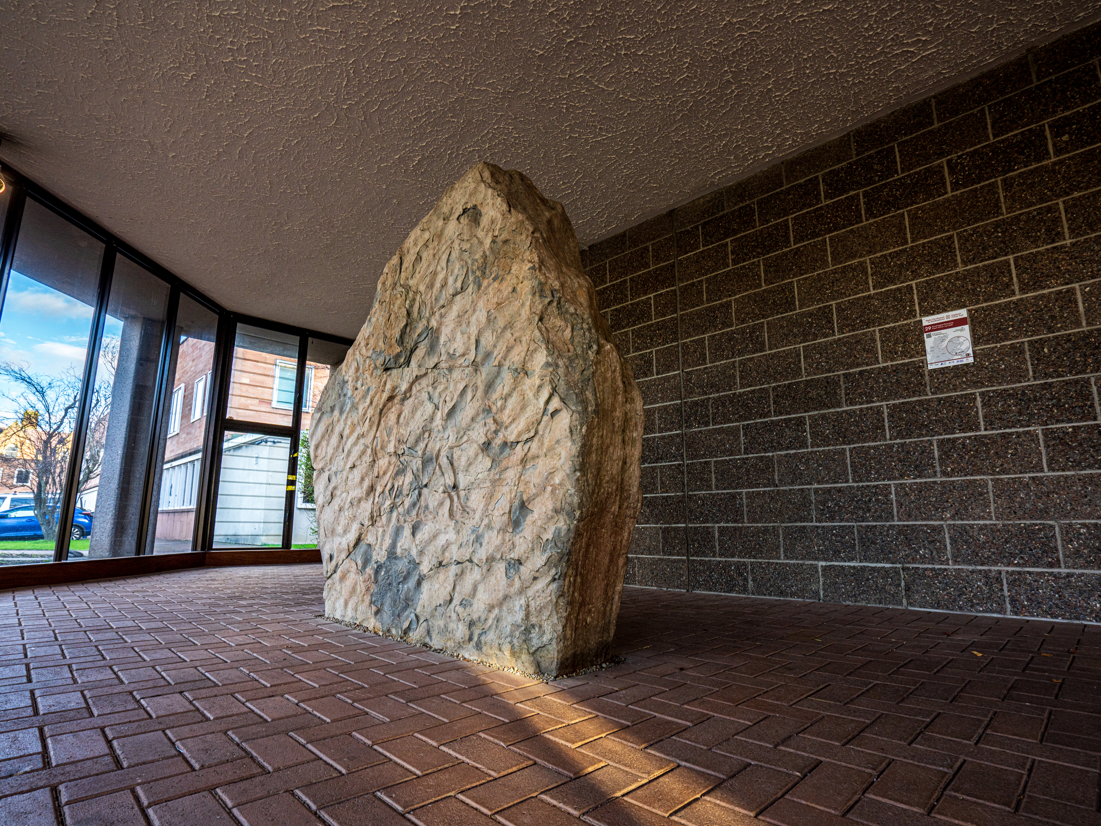 A slightly side on view of the stone displayed indoors on a brick floor, surrounded by glass walls with views of houses and trees outside. The stone is rough and upright, positioned centrally in the room.