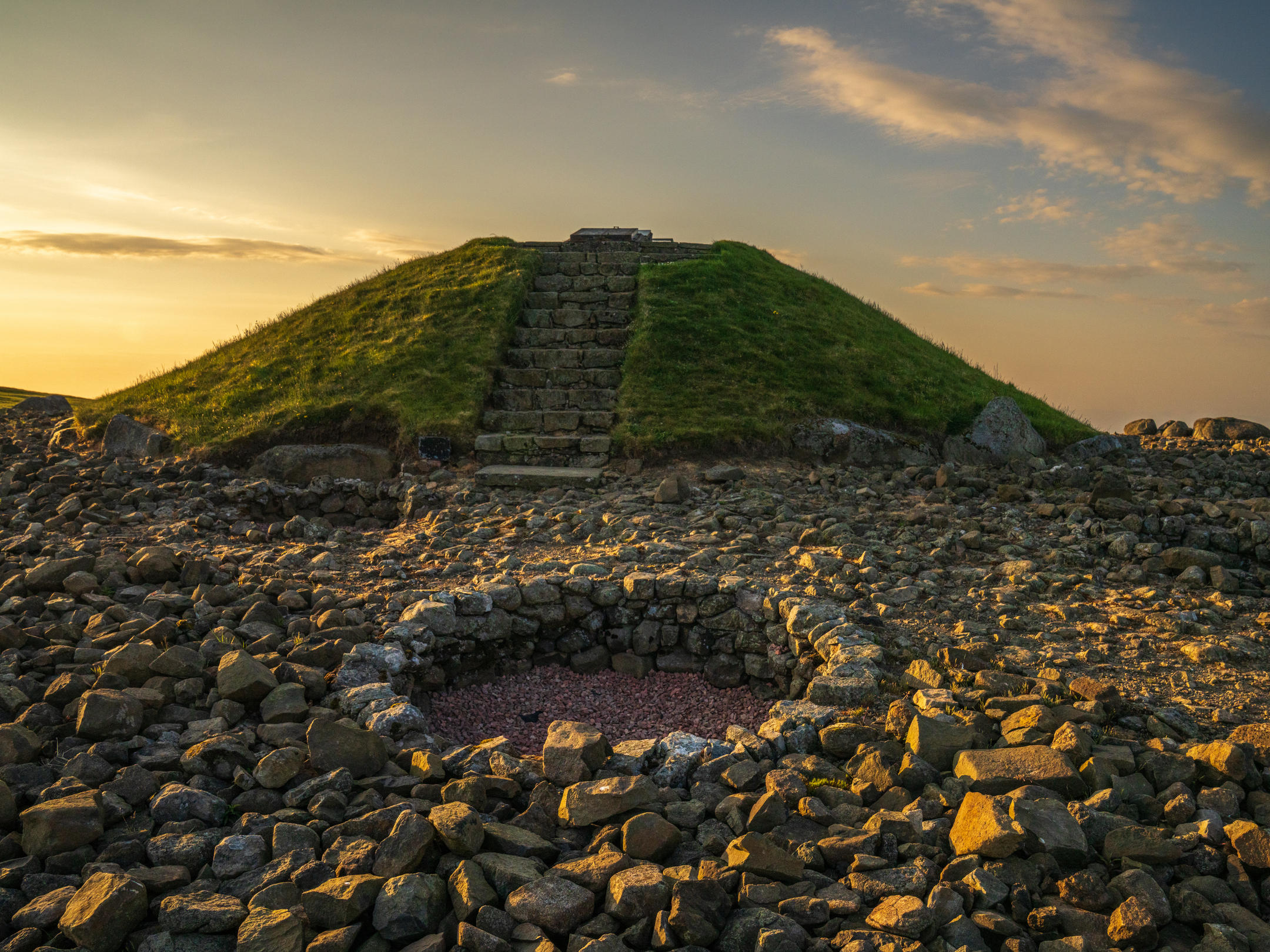 An area of ground covered in stones with holes shaped out of them, and a stairway up a grassy mound in the background