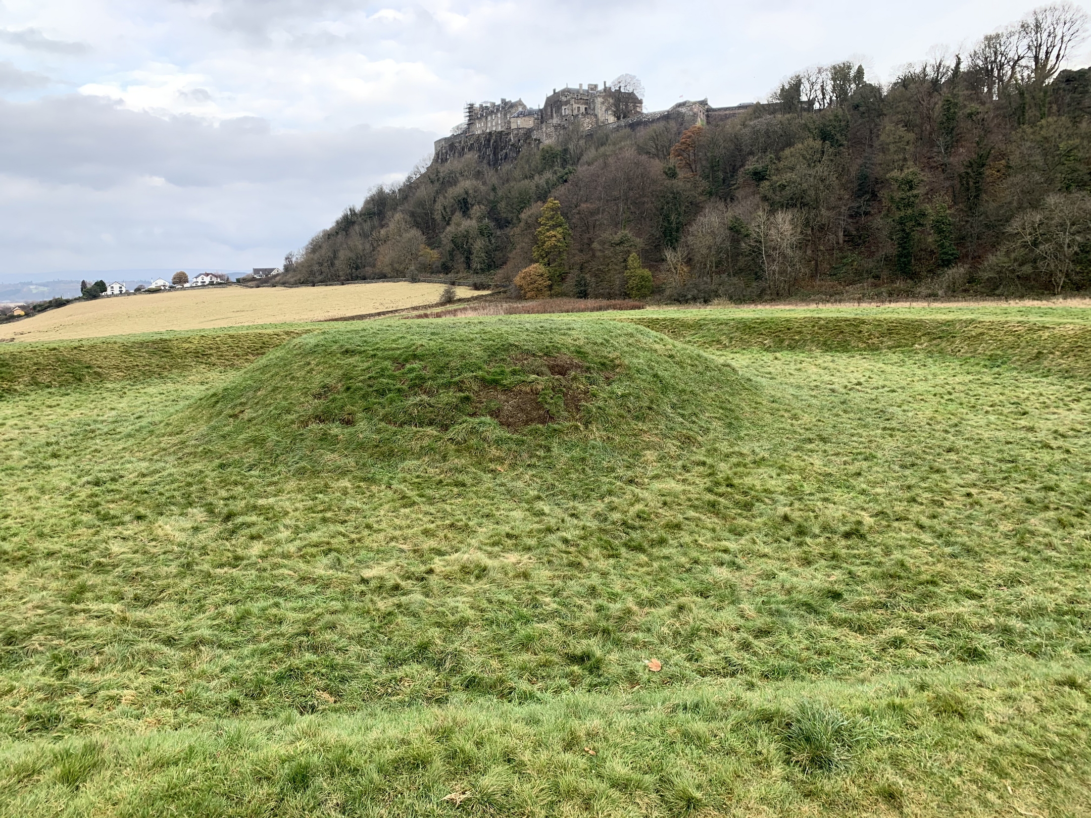 A grassy mound in the King’s Knot area with Stirling Castle rising on a rocky, wooded hill in the background. The castle buildings sit high above autumn-coloured trees under a softly overcast sky.