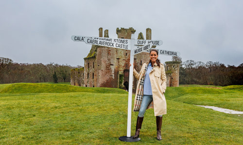 Presenter Jean Johansson stands on a grassy mound in front of Caerlaverock Castle holding onto a post with five signs pointing to heritage sites across Scotland