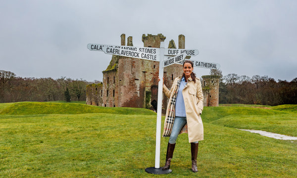 Presenter Jean Johansson stands on a grassy mound in front of Caerlaverock Castle holding onto a post with five signs pointing to heritage sites across Scotland