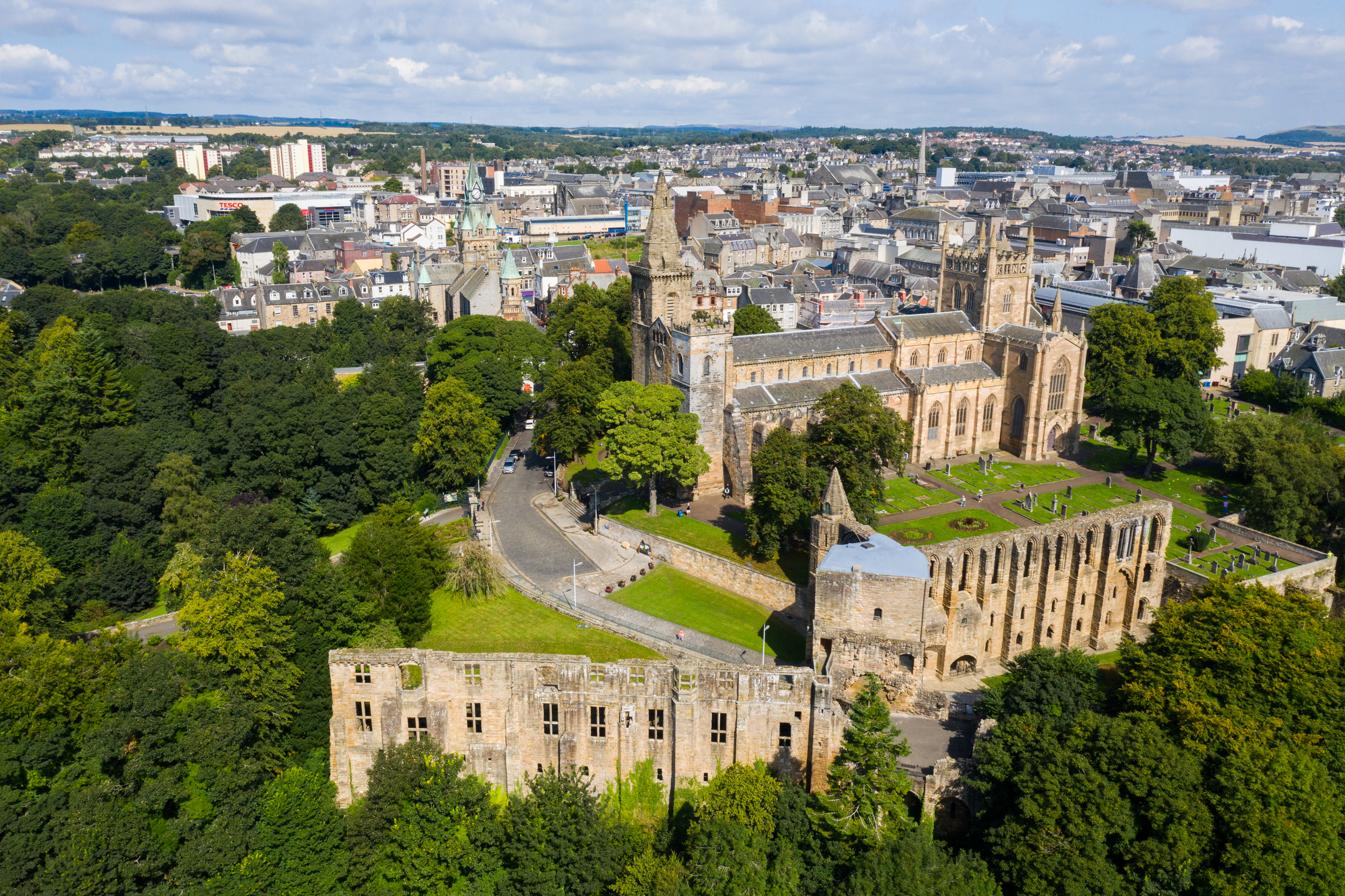 A drone view showing the full expanse of a historic abbey and palace complex, its surrounding trees and graveyard, and nearby town buildings.