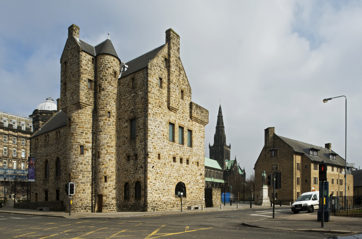 Stone-built St Mungo Museum of Religious Life and Art on a street corner in Glasgow, with its tall, castle‑like towers and small windows. Nearby buildings and the spire of Glasgow Cathedral are visible in the background under a cloudy sky.