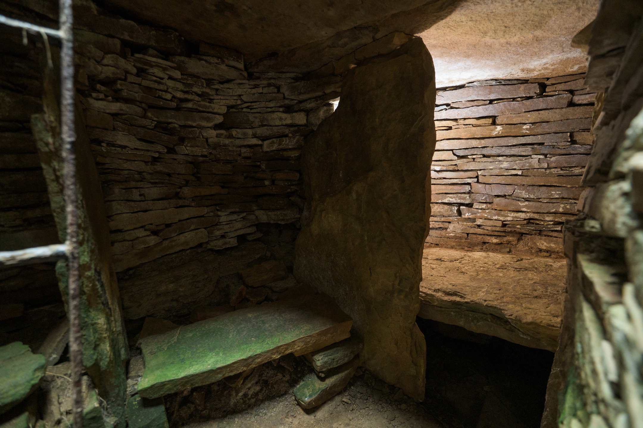 Inside a dark cairn with ladder on the left and stone walls with light in chamber in the distance