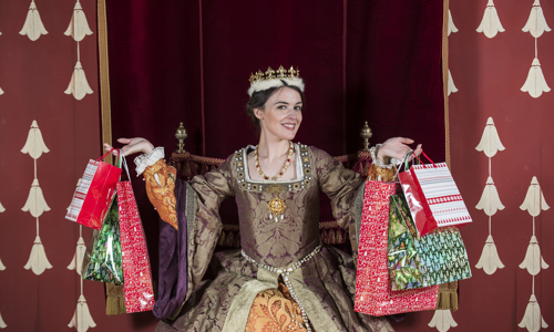 A woman wearing a historical costume of a brown, gold and orange dress with pearl beading sits on a throne. She smiles at the camera and wears a gold crown. In her hands she holds multiple Christmas shopping bags. Behind her is a red velvet curtain.