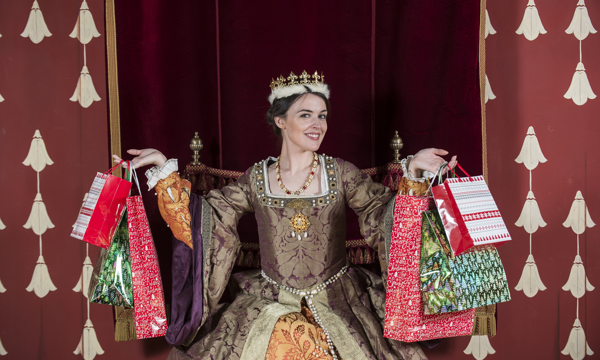 A woman wearing a historical costume of a brown, gold and orange dress with pearl beading sits on a throne. She smiles at the camera and wears a gold crown. In her hands she holds multiple Christmas shopping bags. Behind her is a red velvet curtain.