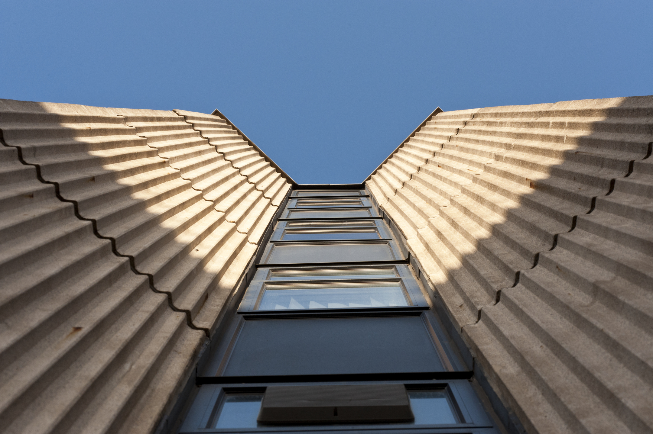 Low‑angle view looking up between two textured concrete walls toward a vertical strip of windows, with a clear blue sky above.