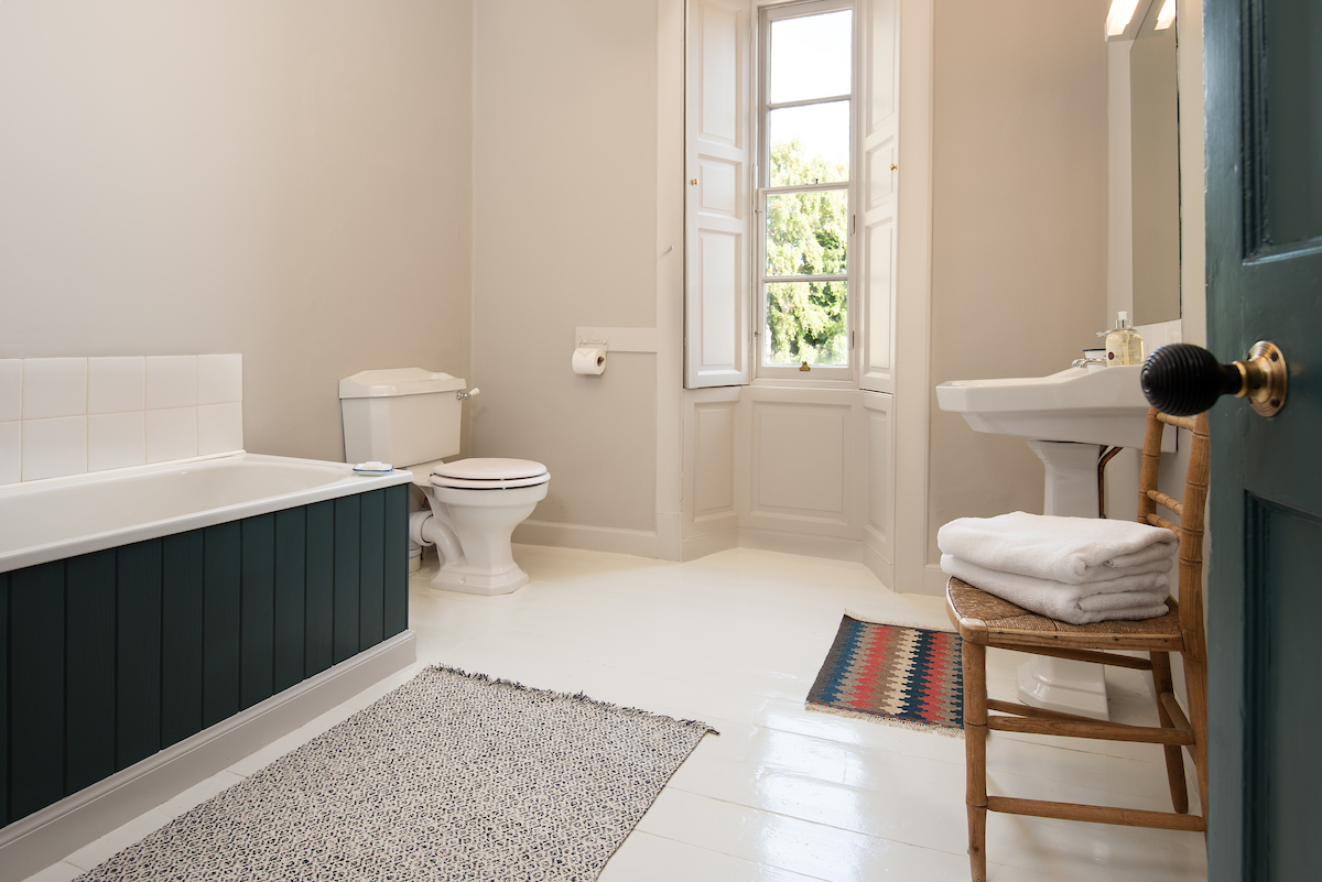 A large bathroom with a white floor and soft grey walls, a wood panelled bath and a narrow sash and case window looking out towards trees