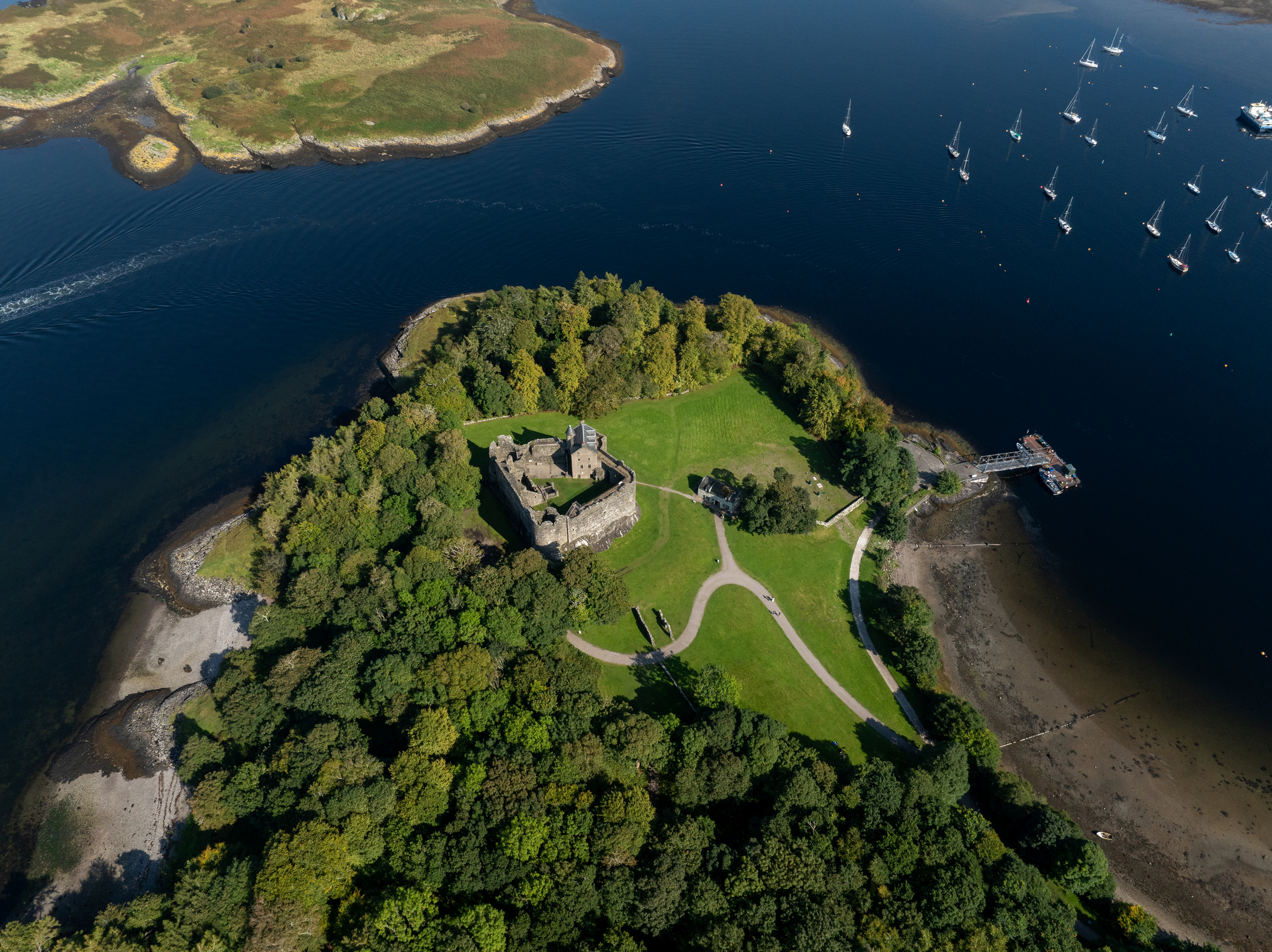 Aerial view of a wooded peninsula featuring a castle at its centre, surrounded by water and moored boats.