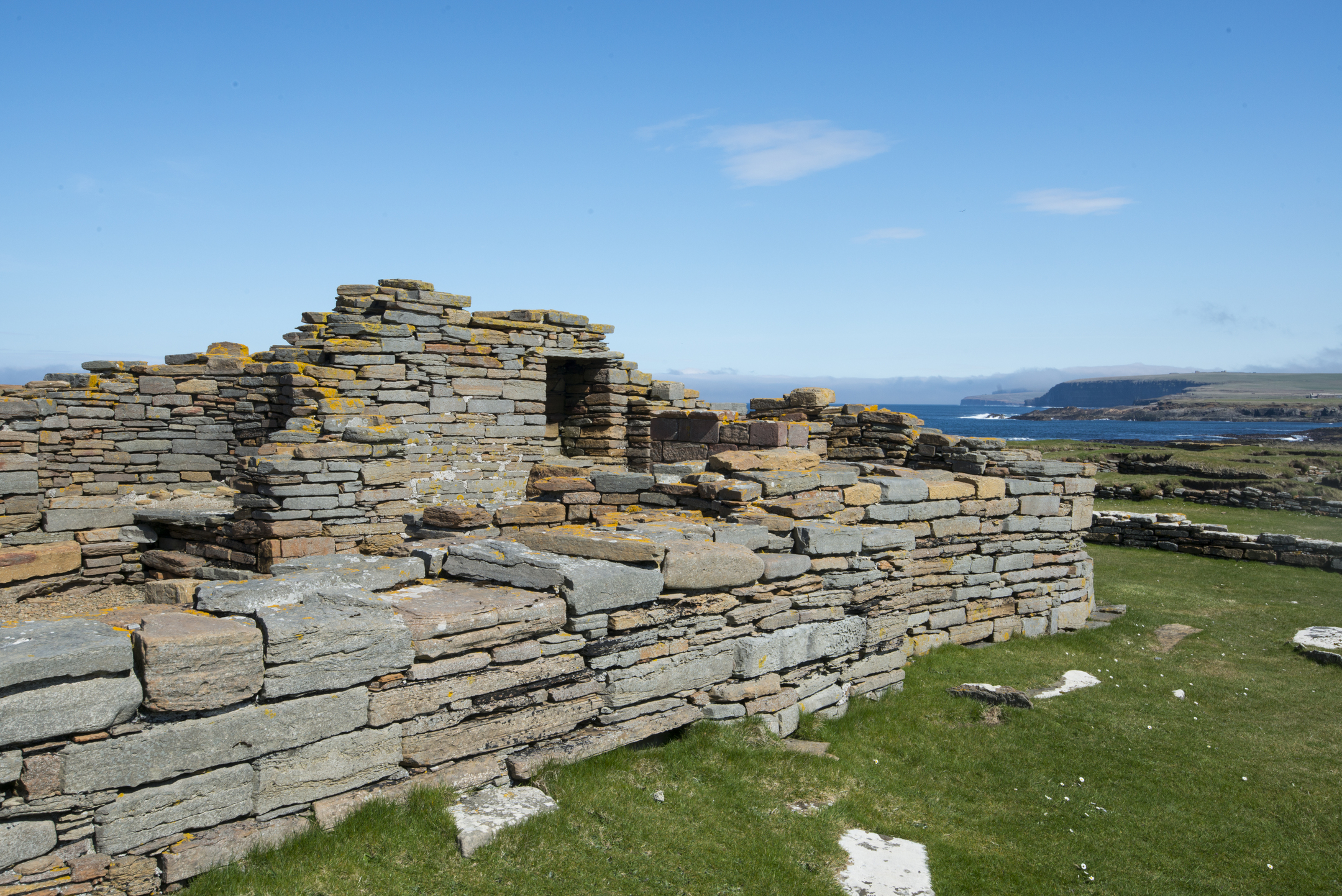 A partially standing stone wall with layered stone construction, with the blue sea and distant cliffs visible behind it.