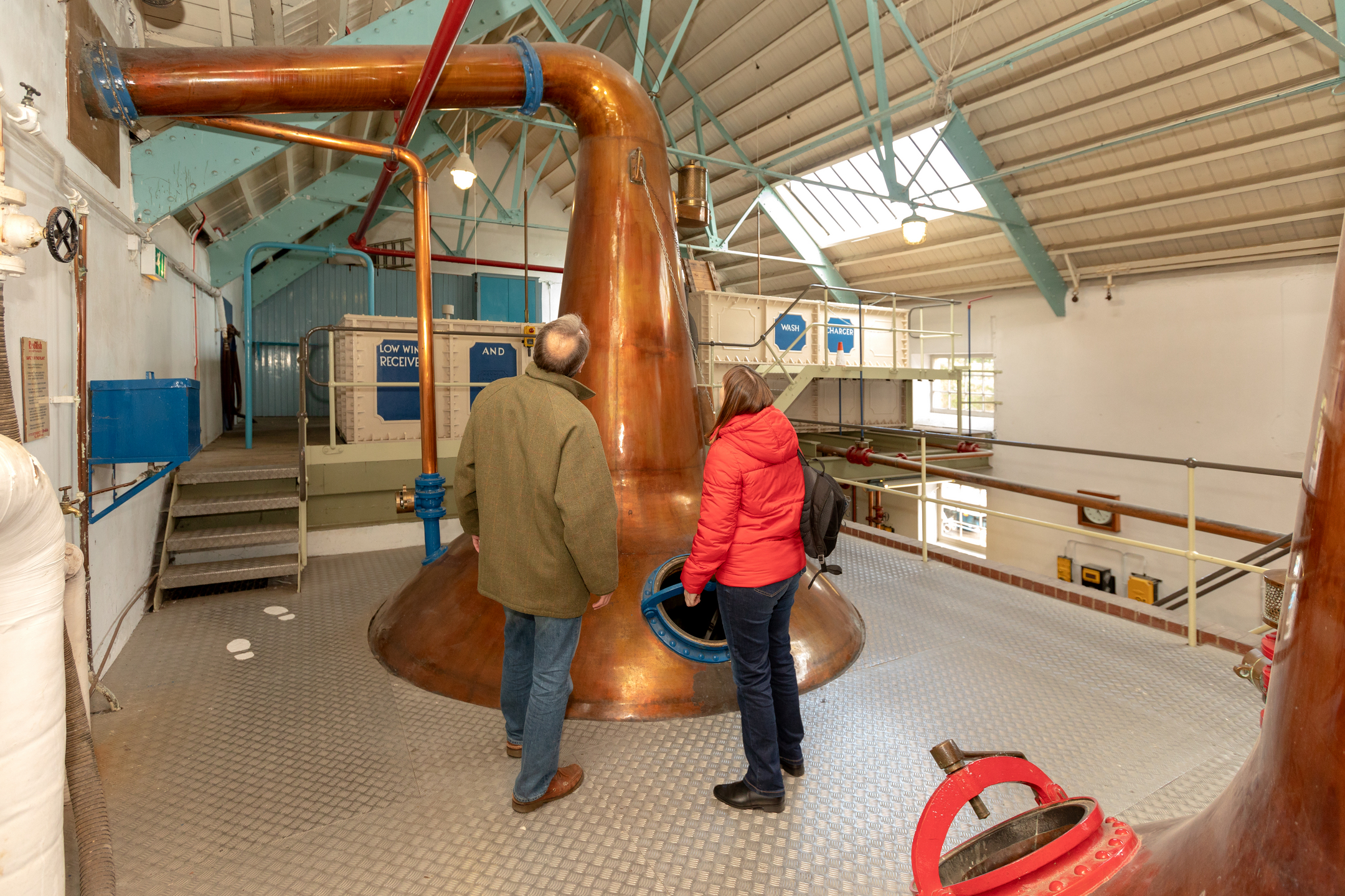Visitors stand beside a large copper pot still inside the historic Dallas Dhu distillery, surrounded by traditional distilling equipment, pipework and industrial walkways.