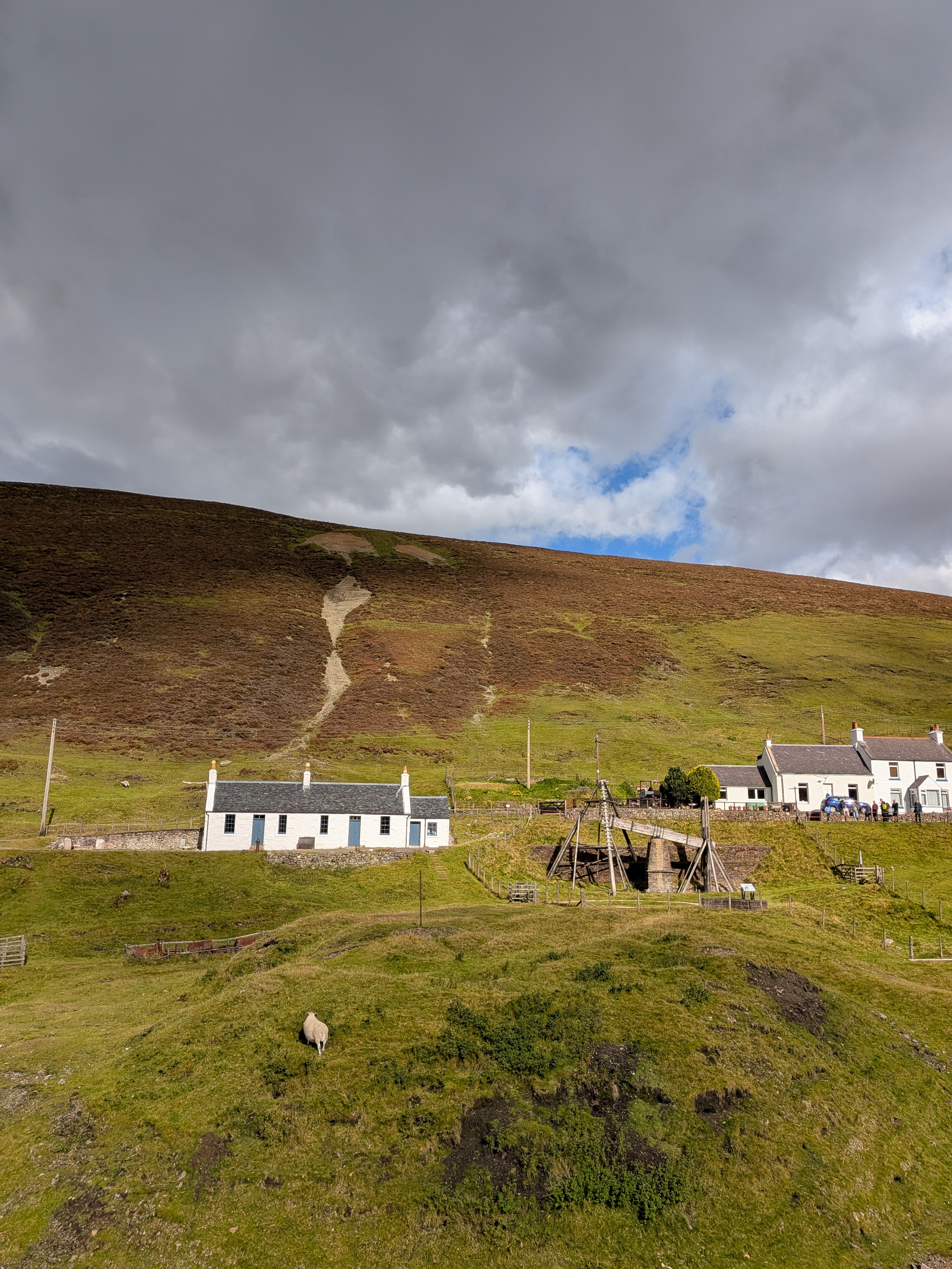 A wooden pump, a relic from the lead mining industry, in front of a row of quaint whitewashed cottages. Behind the buildings there is an expanse of barren moorland.