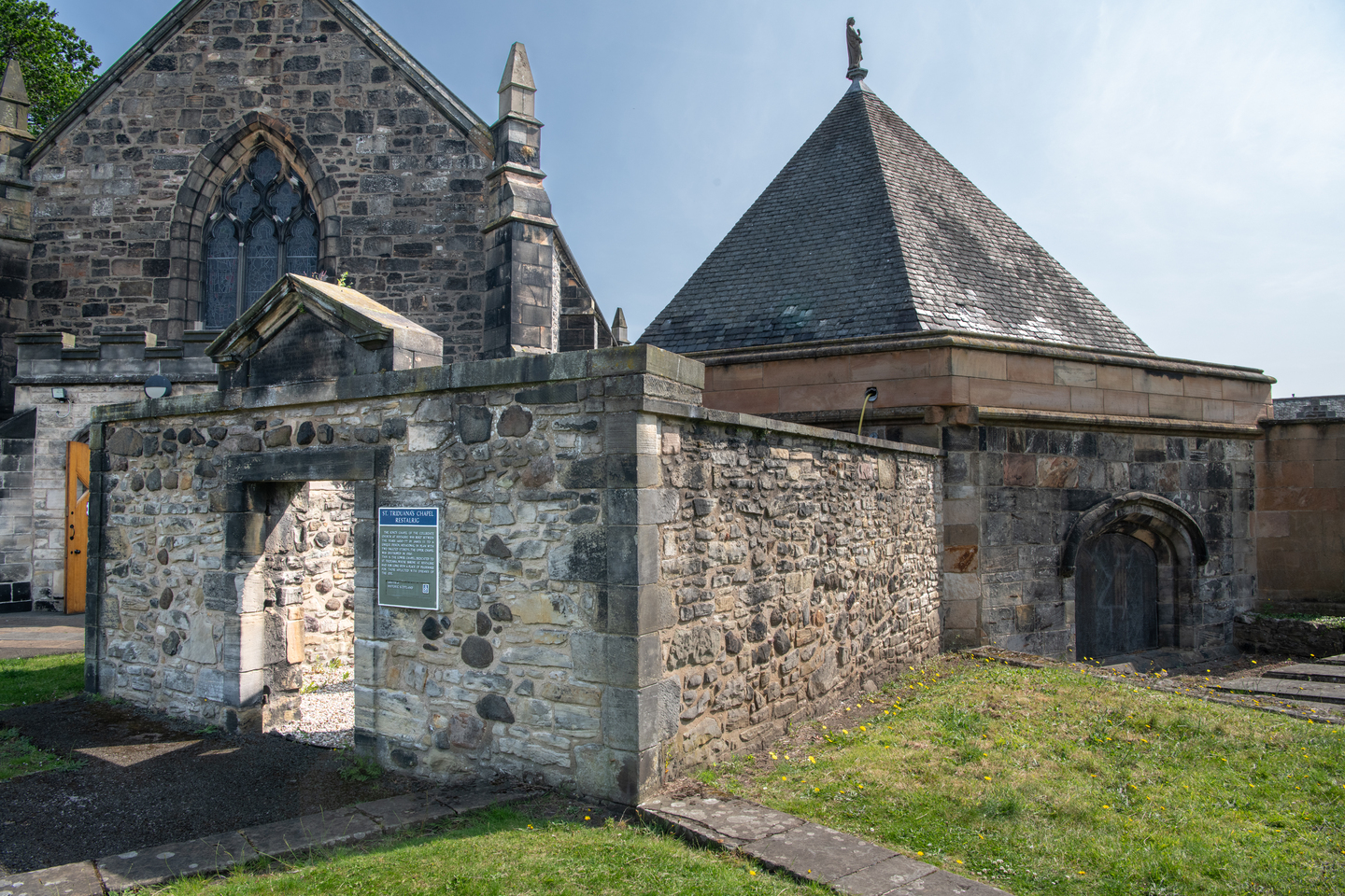 The side of a collection of stone buildings, in various shapes. The front building is in a short, square shape, with an entrance in the middle with light coming through. Behind are two taller buildings, one resembling a religious building.  There is grass in front of them. 