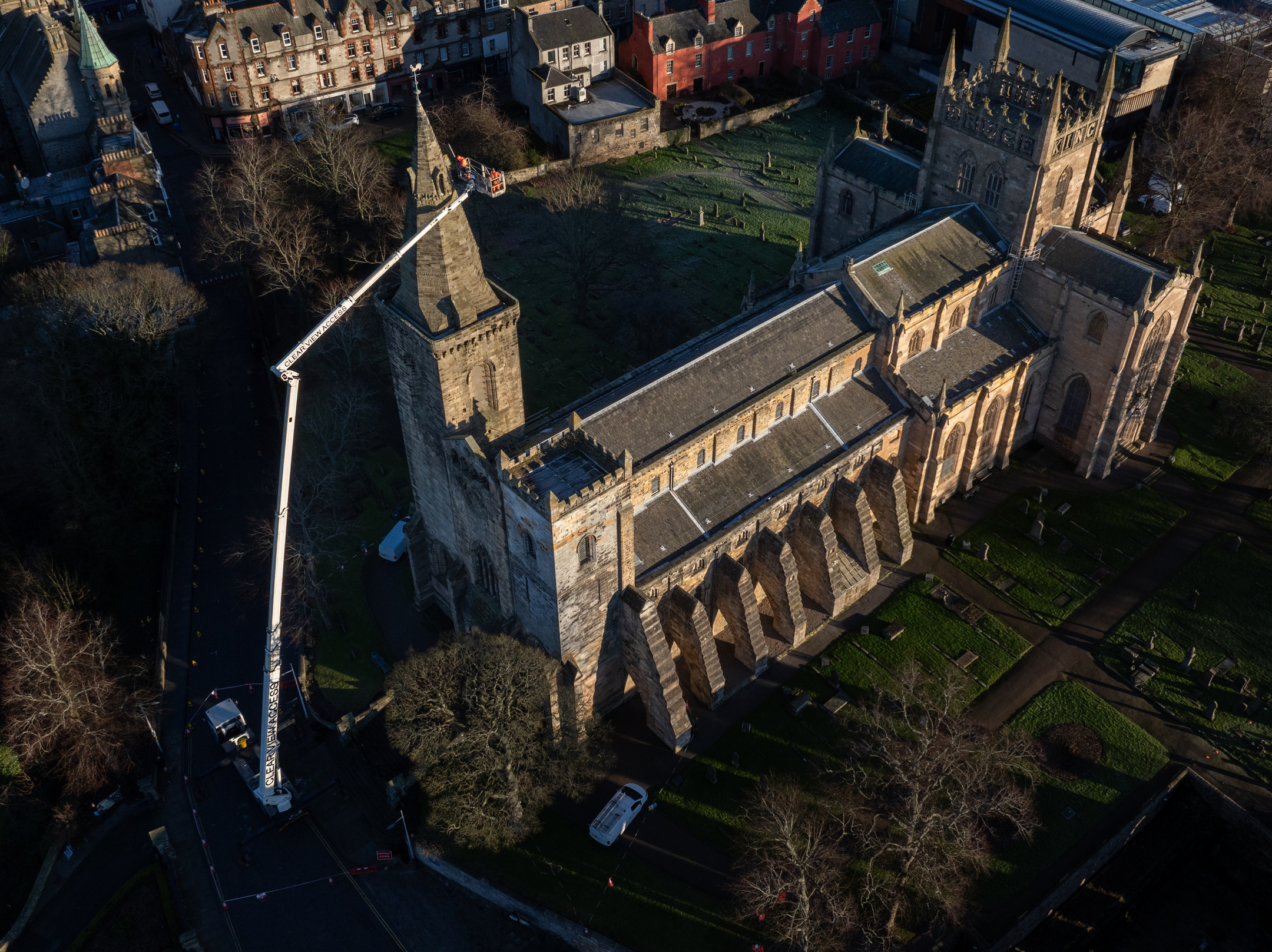 A high-angle drone view of a large stone abbey, with a tall lift raised to the tower for external inspection work. Surrounding streets, trees, and nearby buildings are visible.