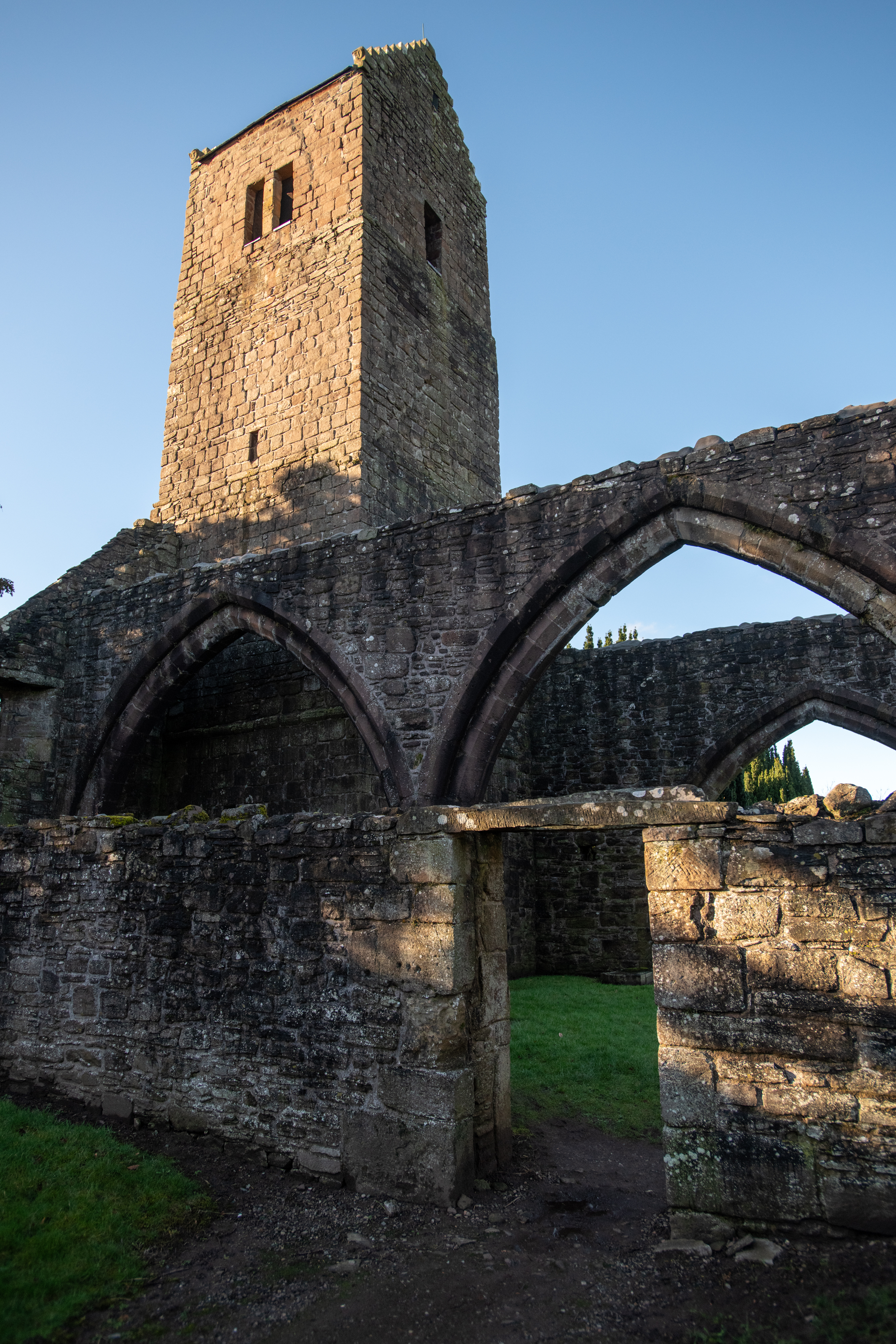 A close view of the church tower rising above the surviving arched walls, with low sunlight casting shadows across the stonework and grass.