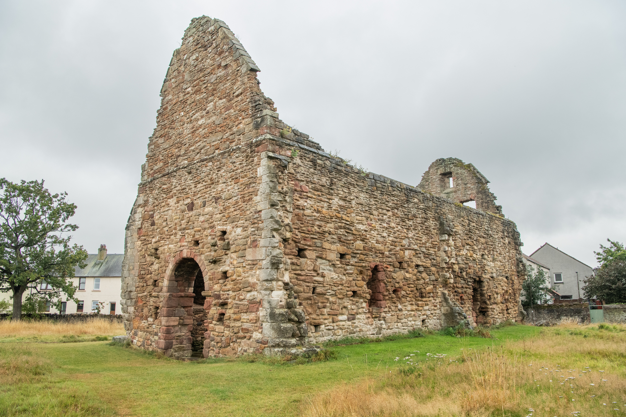 An angled exterior view of an old stone church ruin surrounded by green grass, with houses visible behind it.