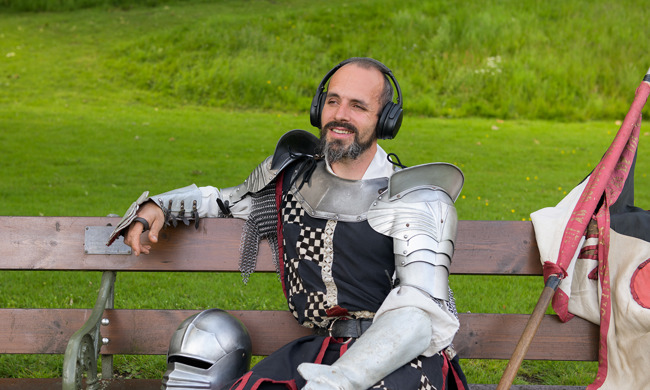 A performer dressed as a knight is sitting in a park bench with headphones on his head. He is smiling and looking relaxed. 