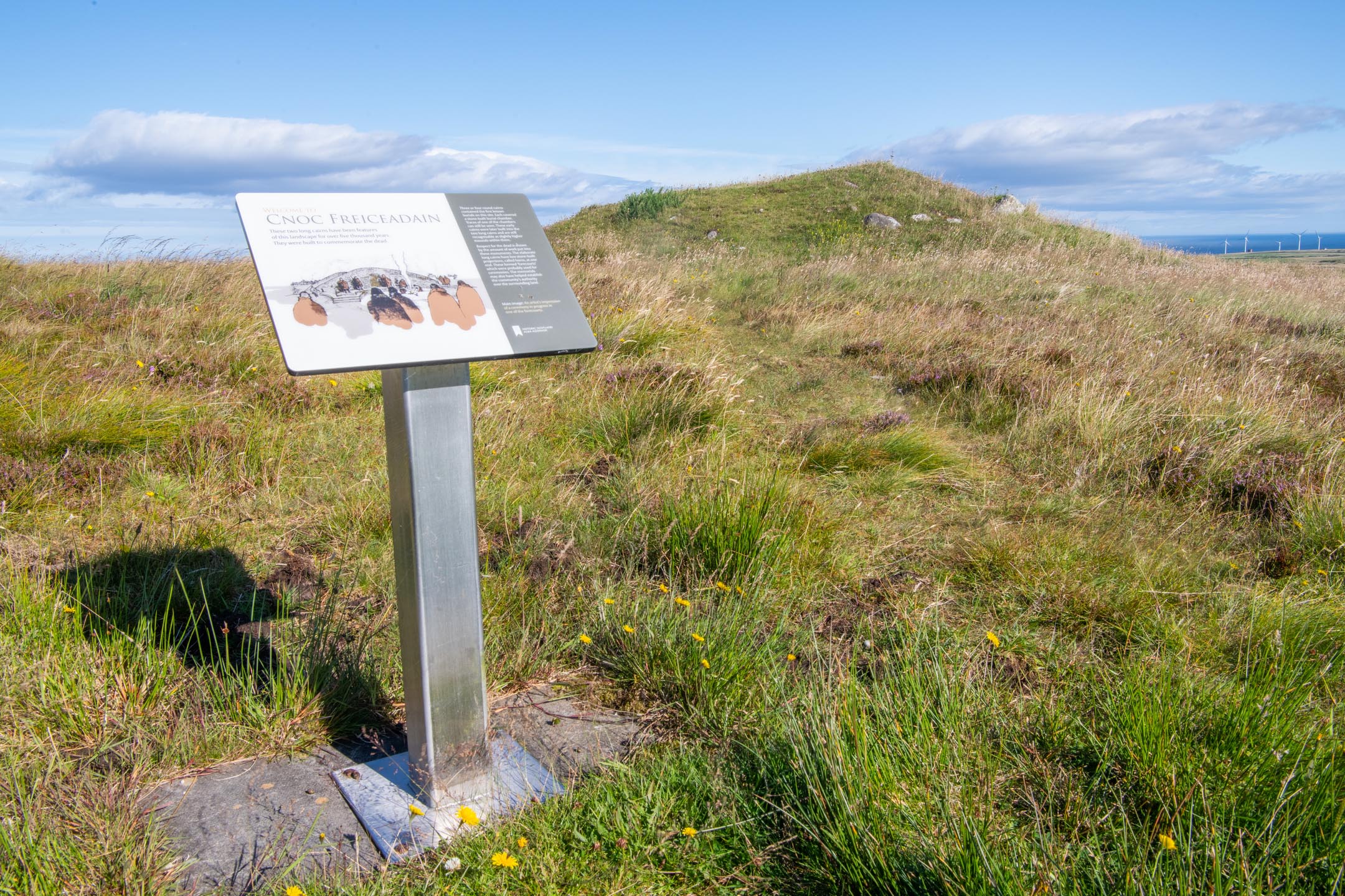 Information panel in front of grassy cairn with blue sky and distant sea.