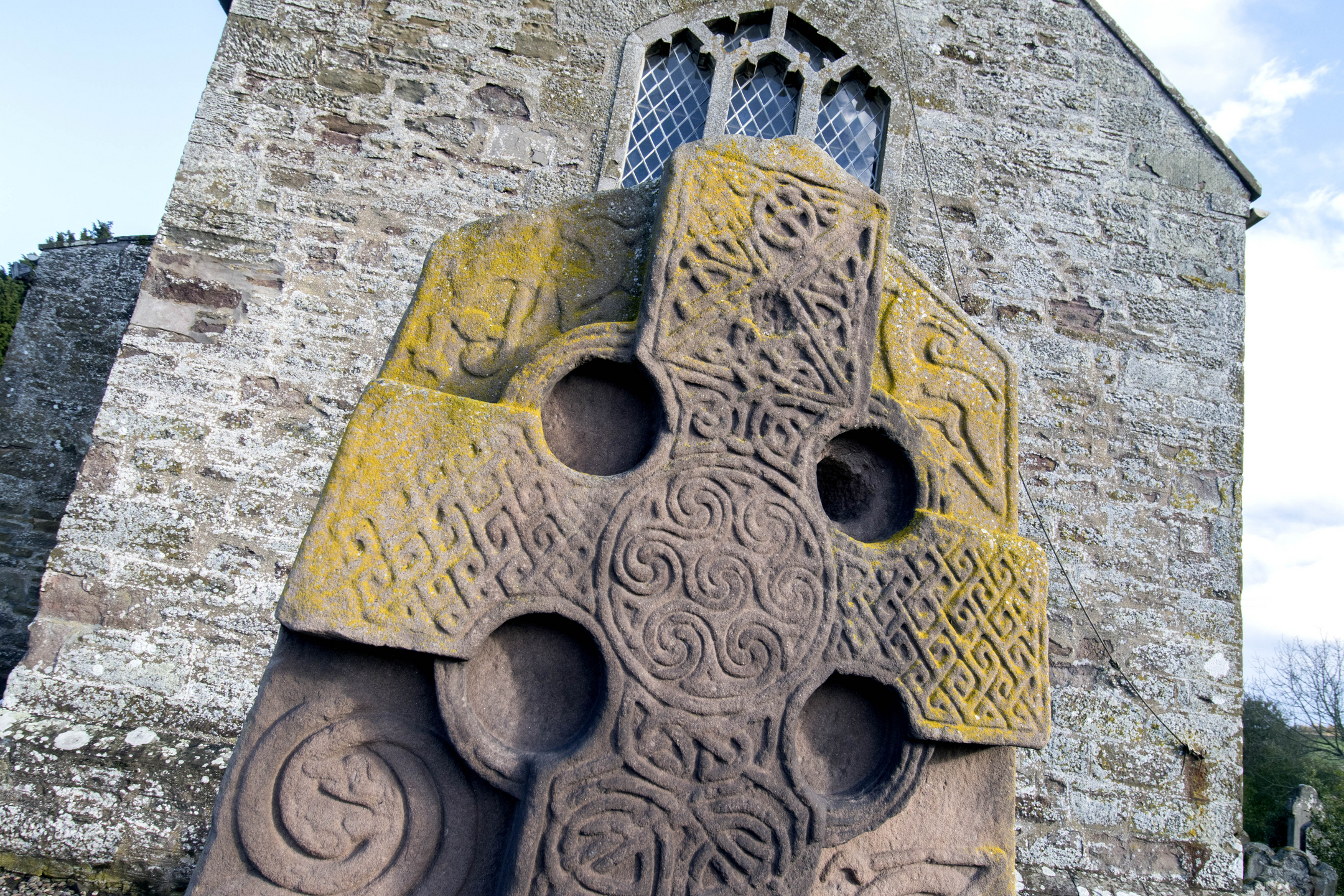 Close-up at an angle of the top of a tall, cross-shaped sculptured stone with yellow discolouration