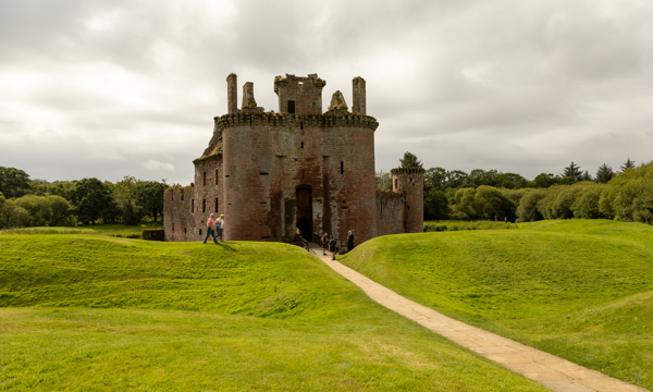 A stone castle with a moat surrounded by grass with a path leading from the entrance. There is also a bridge at the entrance. The castle is red in colour and a triangular shape and there is grass on either side of the path. The castle has many window gaps in the walls and there are two circular towers at the front and one at the back right-hand side. The sky is cloudy and there are people walking on the grass and a group of people standing at the end of the bridge near the entrance.