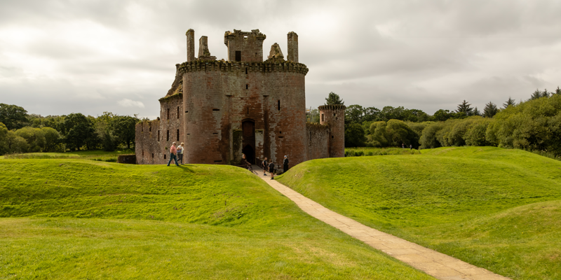 A stone castle with a moat surrounded by grass with a path leading from the entrance. There is also a bridge at the entrance. The castle is red in colour and a triangular shape and there is grass on either side of the path. The castle has many window gaps in the walls and there are two circular towers at the front and one at the back right-hand side. The sky is cloudy and there are people walking on the grass and a group of people standing at the end of the bridge near the entrance.