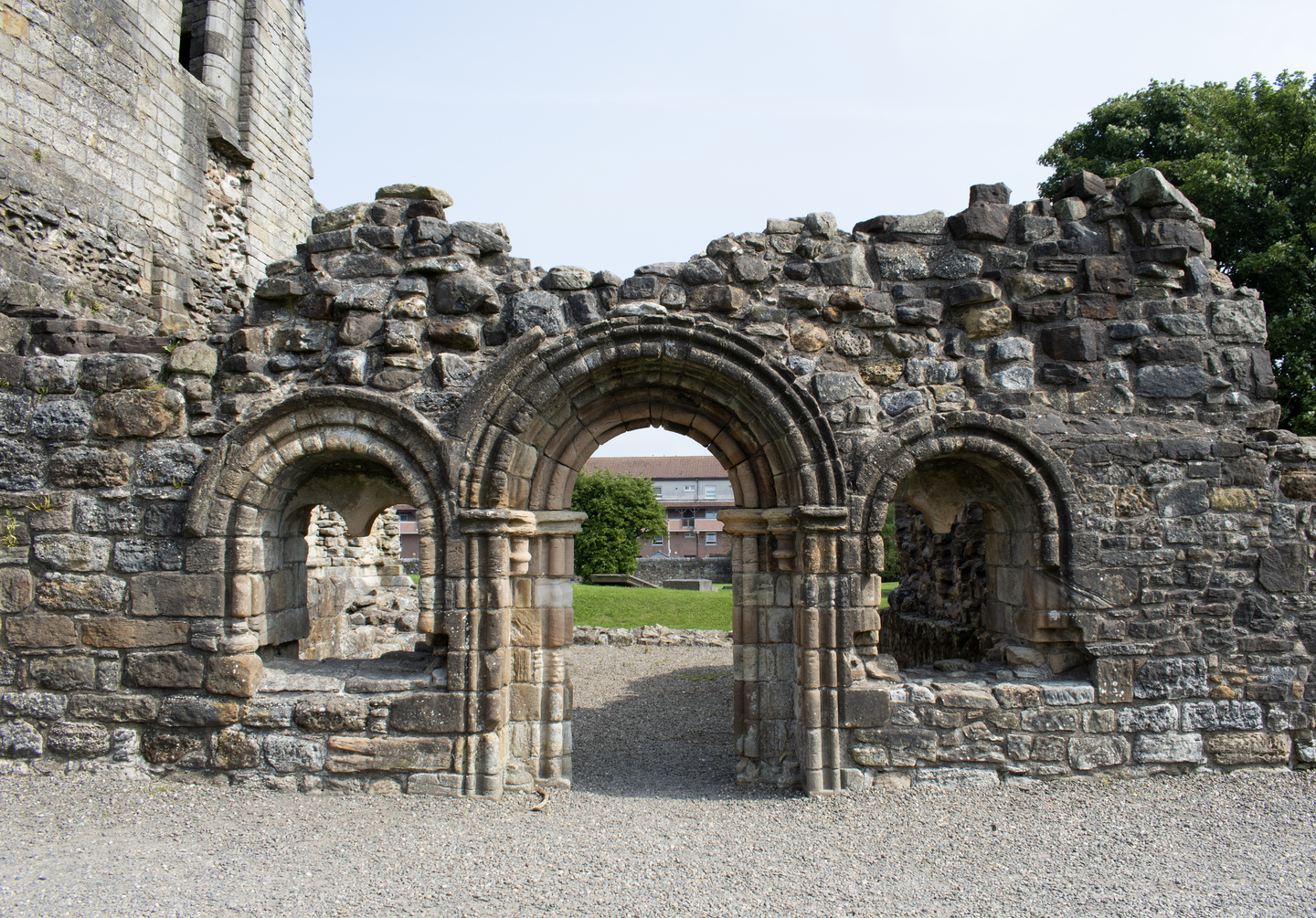 Section of a ruined stone wall featuring three rounded arches. The central arch forms a doorway opening onto grass and modern buildings in the distance. The wall is built from uneven stones, showing signs of age