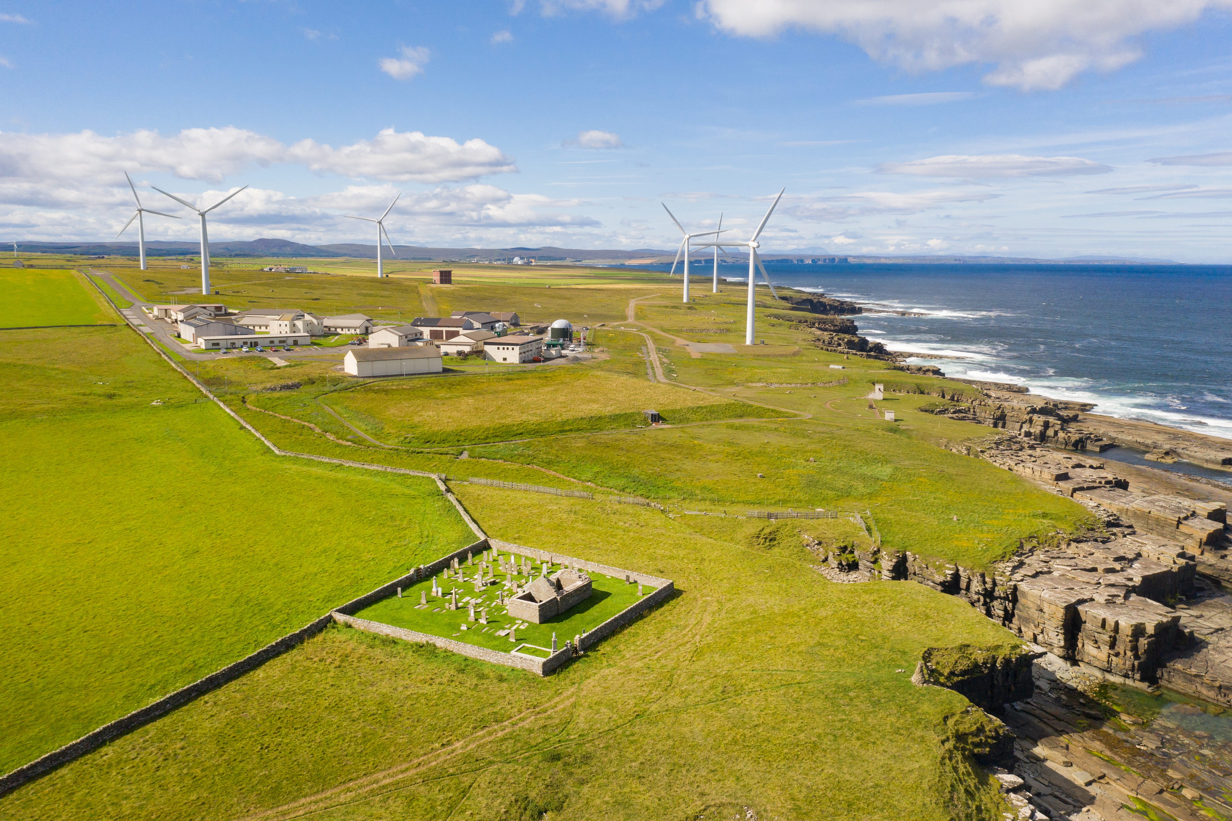 An aerial photo showing the ruins of a historic chapel in the foreground and a modern business park and wind farm in the background. All are set in a very rural landscape close to dramatic sea cliffs.