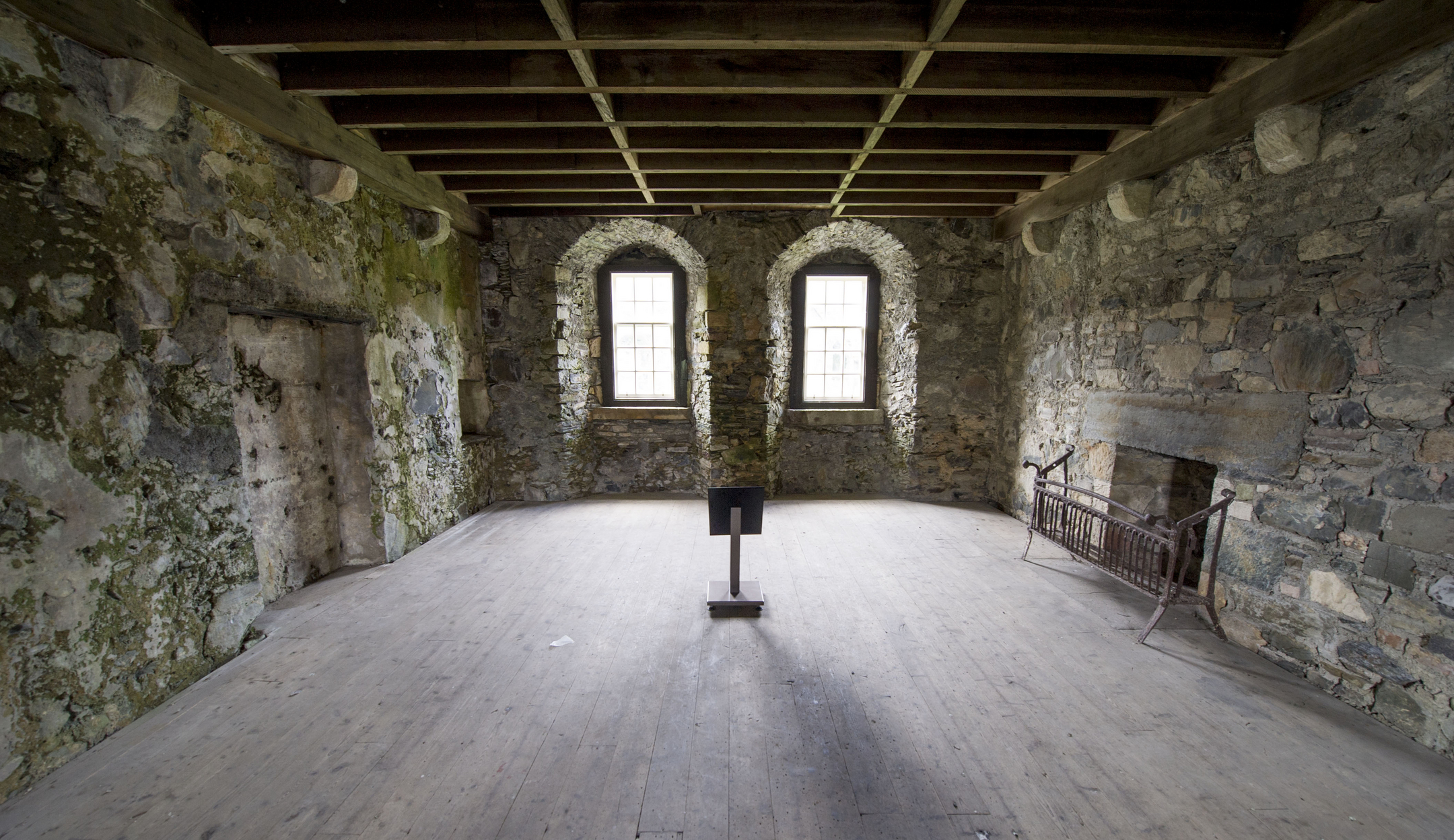 A rectangular stone room with three arched windows along the back wall, a large stone fireplace on the right, and a wooden beam ceiling above, with a display stand positioned in the middle of the floor.